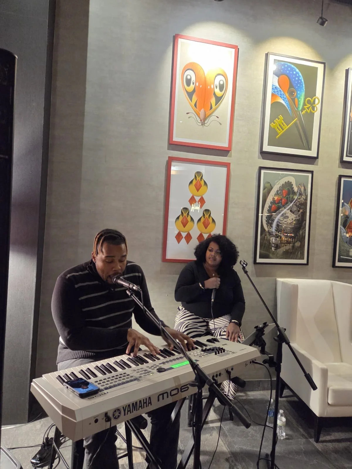 Musician playing a Yamaha keyboard with a woman singing behind him in a modern art decorated room.