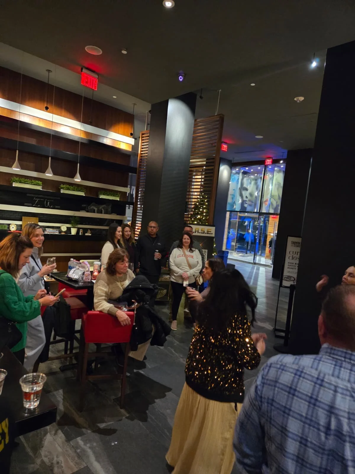 Group of people gathered in a modern indoor venue, some holding drinks, with a Christmas tree and decorative elements in the background.