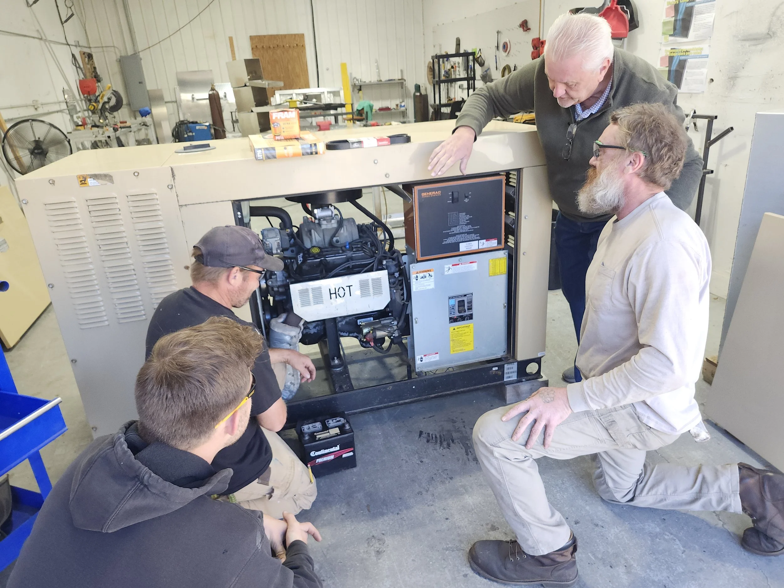 Four men working on a large industrial generator or compressor inside a workshop. One man is kneeling and working on the machine, while the other three observe and discuss. The machine has a label that reads 'HOT' and other labels and screens on its front panel.