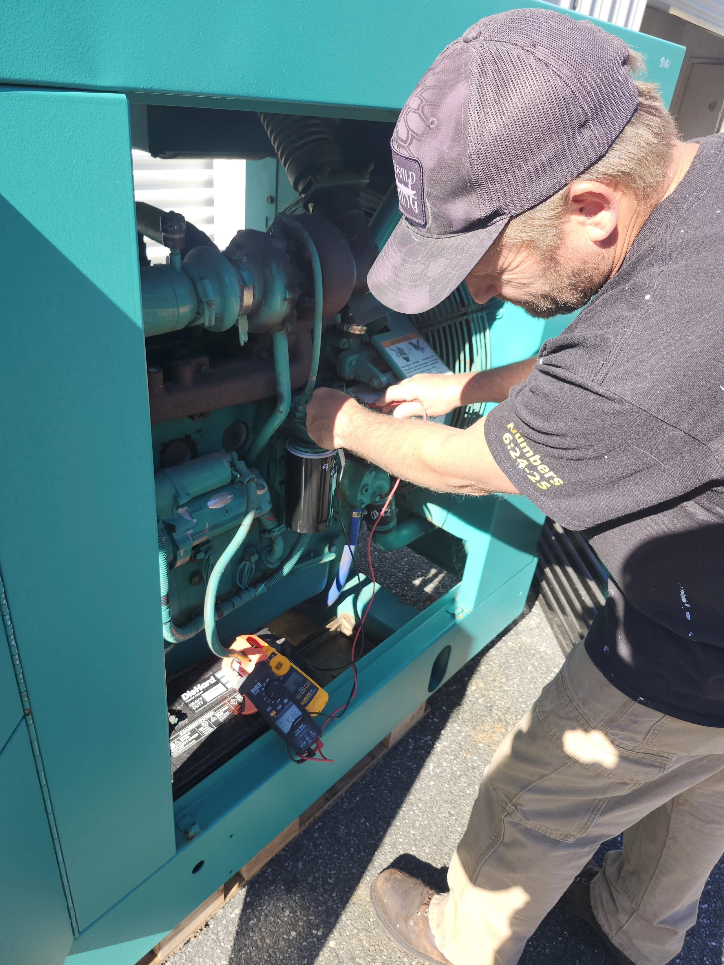 Man wearing a gray cap and black T-shirt working on a teal-colored industrial generator or machinery outside, using a multimeter to measure electrical current.