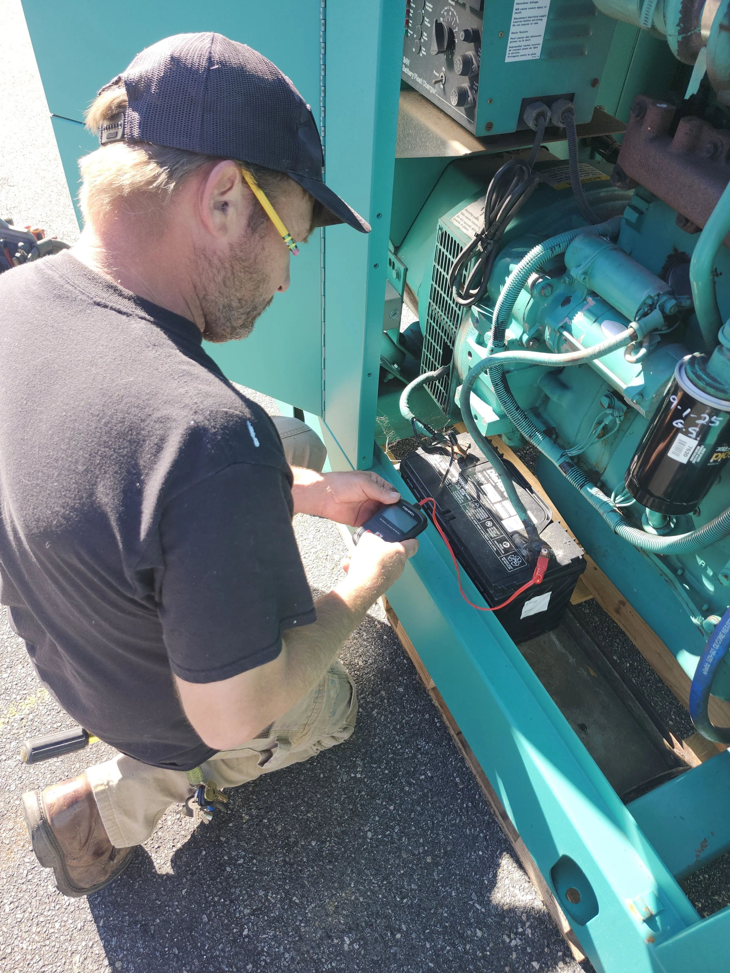 A man working on a generator or engine, using a multimeter to test the battery. He is wearing a black t-shirt, khaki pants, a navy blue cap, and yellow safety glasses.