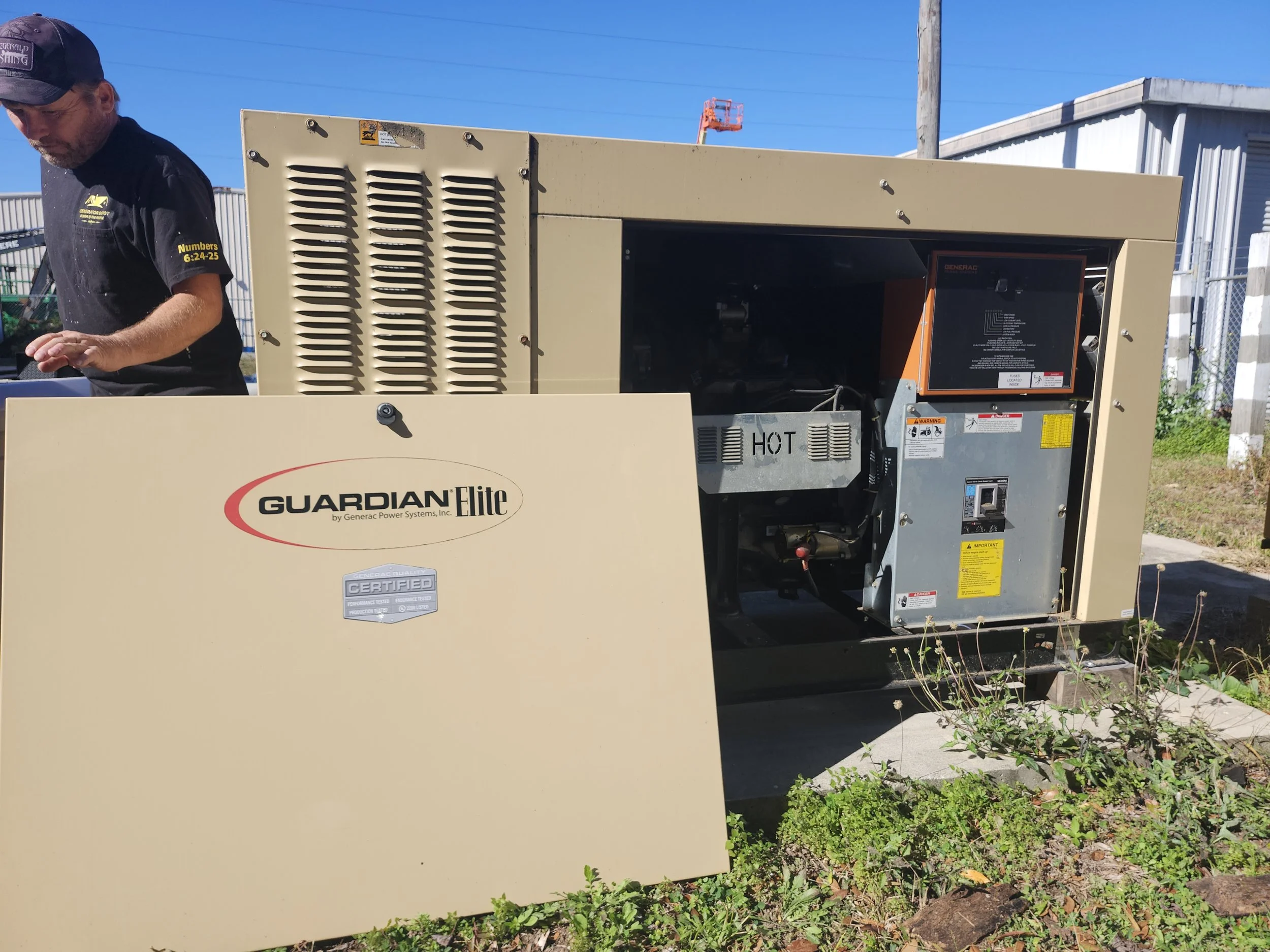 A beige Guardian Elite generator with its side panel removed, revealing internal components. A man is standing to the left, wearing a black shirt and cap, outdoors on a sunny day with a blue sky and some industrial buildings and fencing in the background.