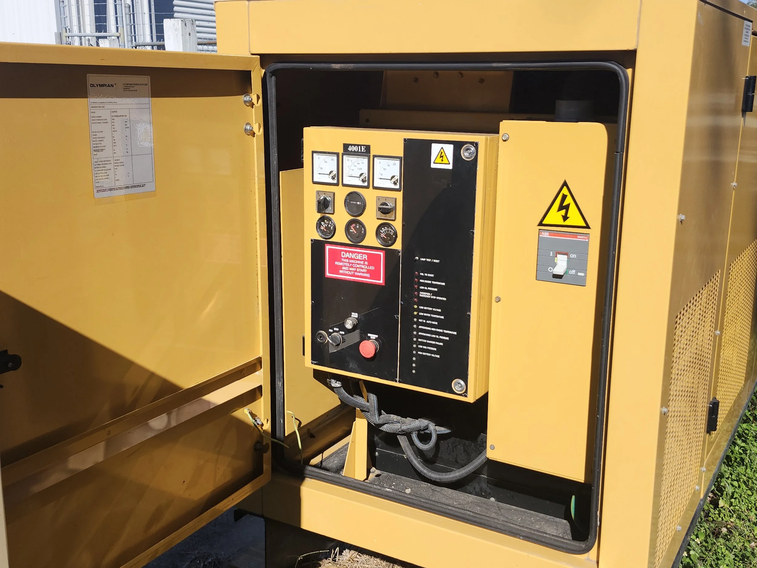 Yellow industrial generator with control panel, warning labels, and circuit breaker, situated outdoors near a grassy area.
