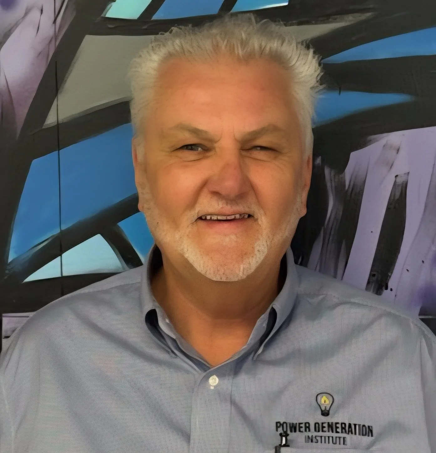 A smiling older man with gray hair and a beard, wearing a gray collared shirt with a Power Generation Institute logo, standing in front of a large model aircraft.