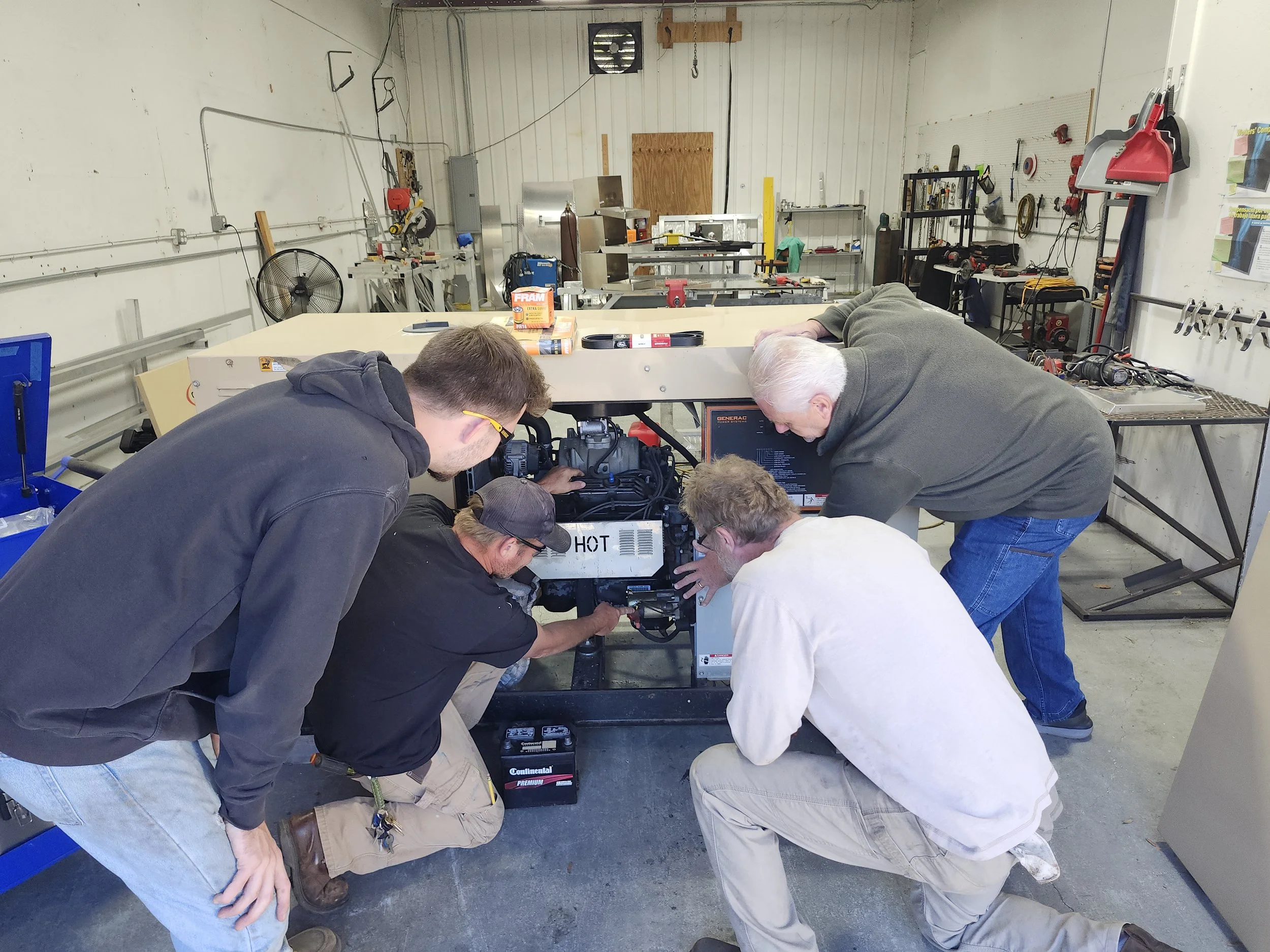 Four men working on a small engine or machine in a workshop or garage, with tools and equipment on the tables and walls around them.