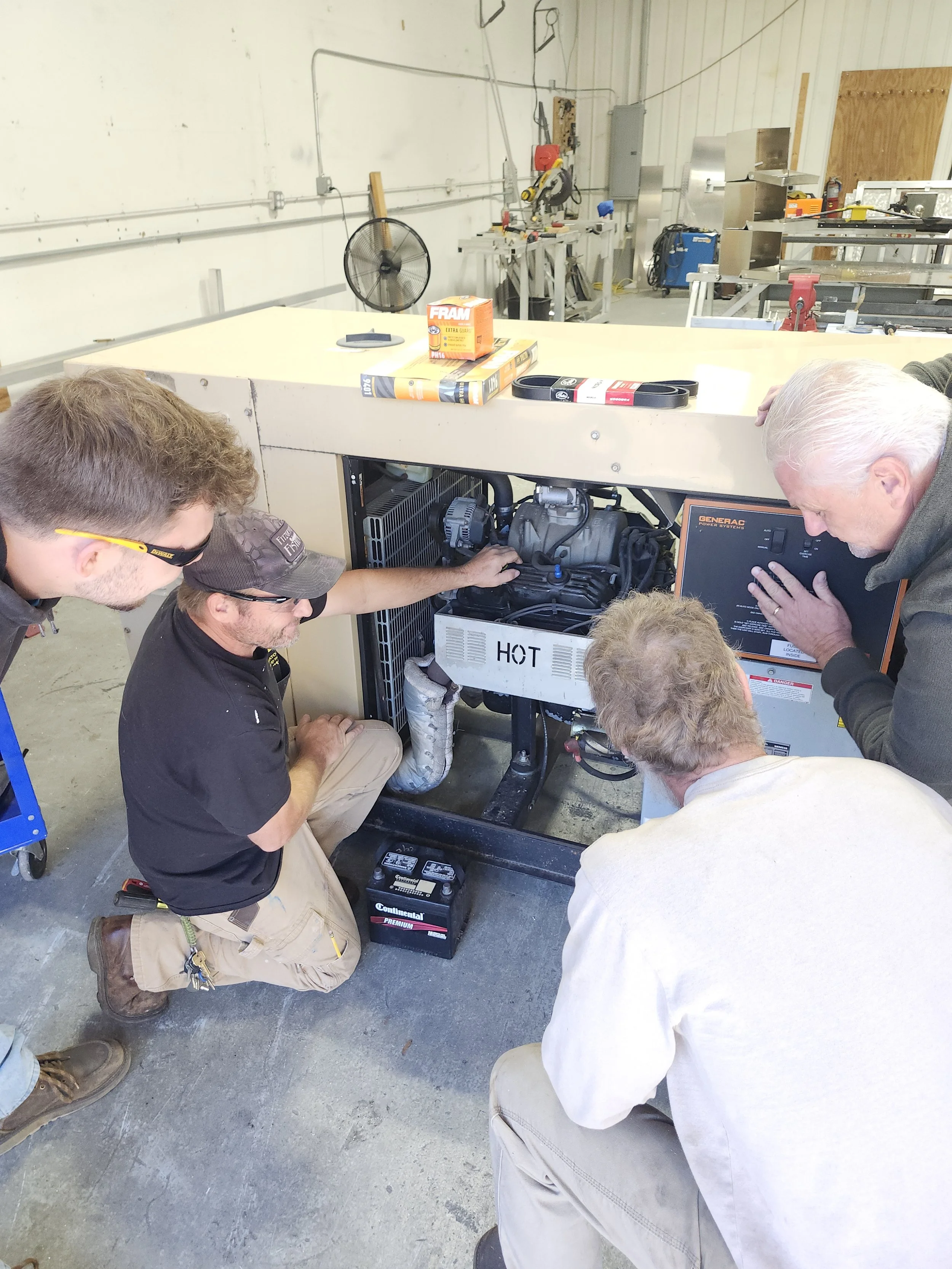 Five men are working on a generator or industrial equipment in a workshop. One man is kneeling, and four men are standing closely observing or discussing the equipment.