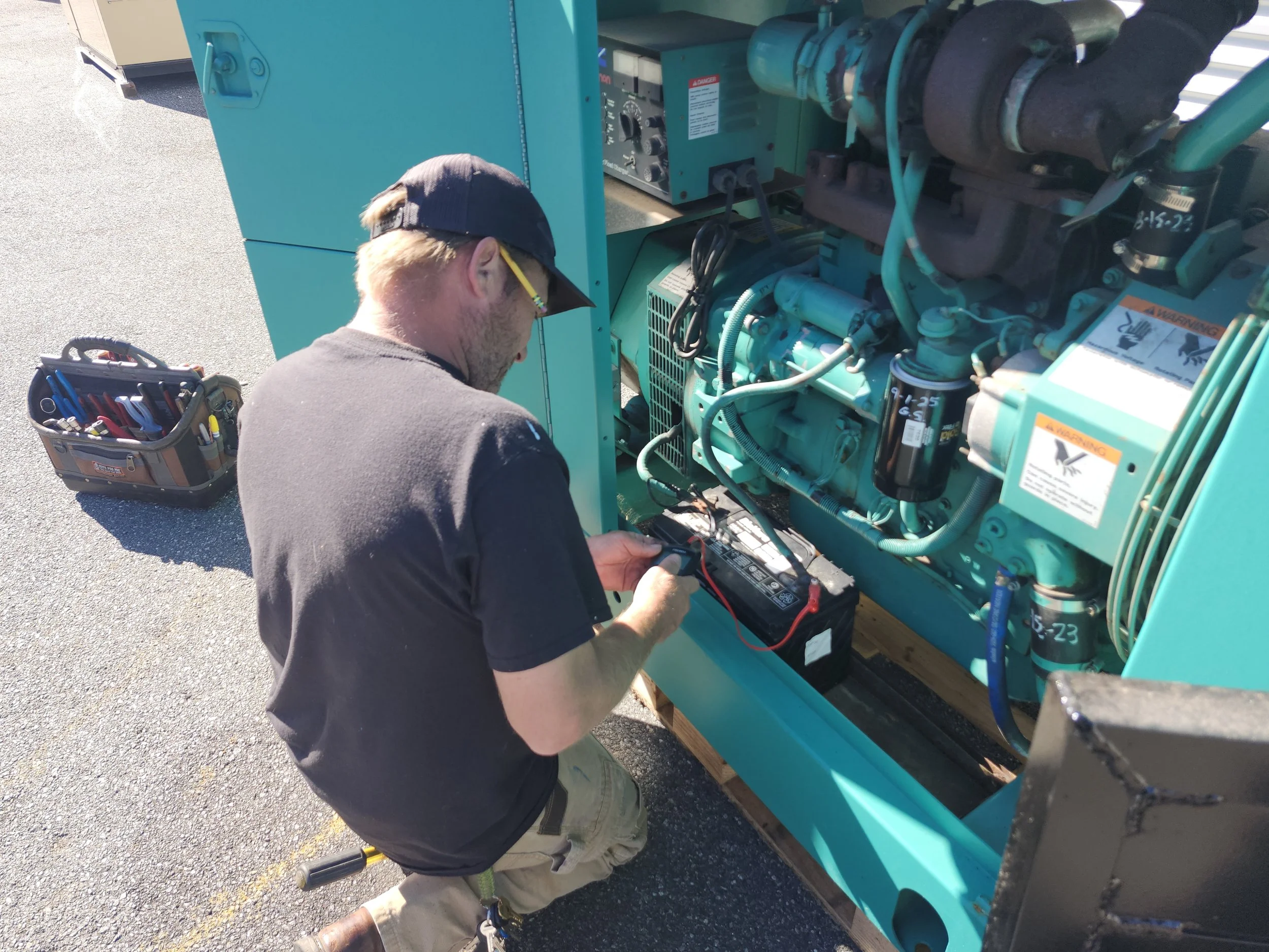 Technician working on a large teal-colored industrial generator or machinery outdoors, using a multimeter and tools from a nearby toolbox.