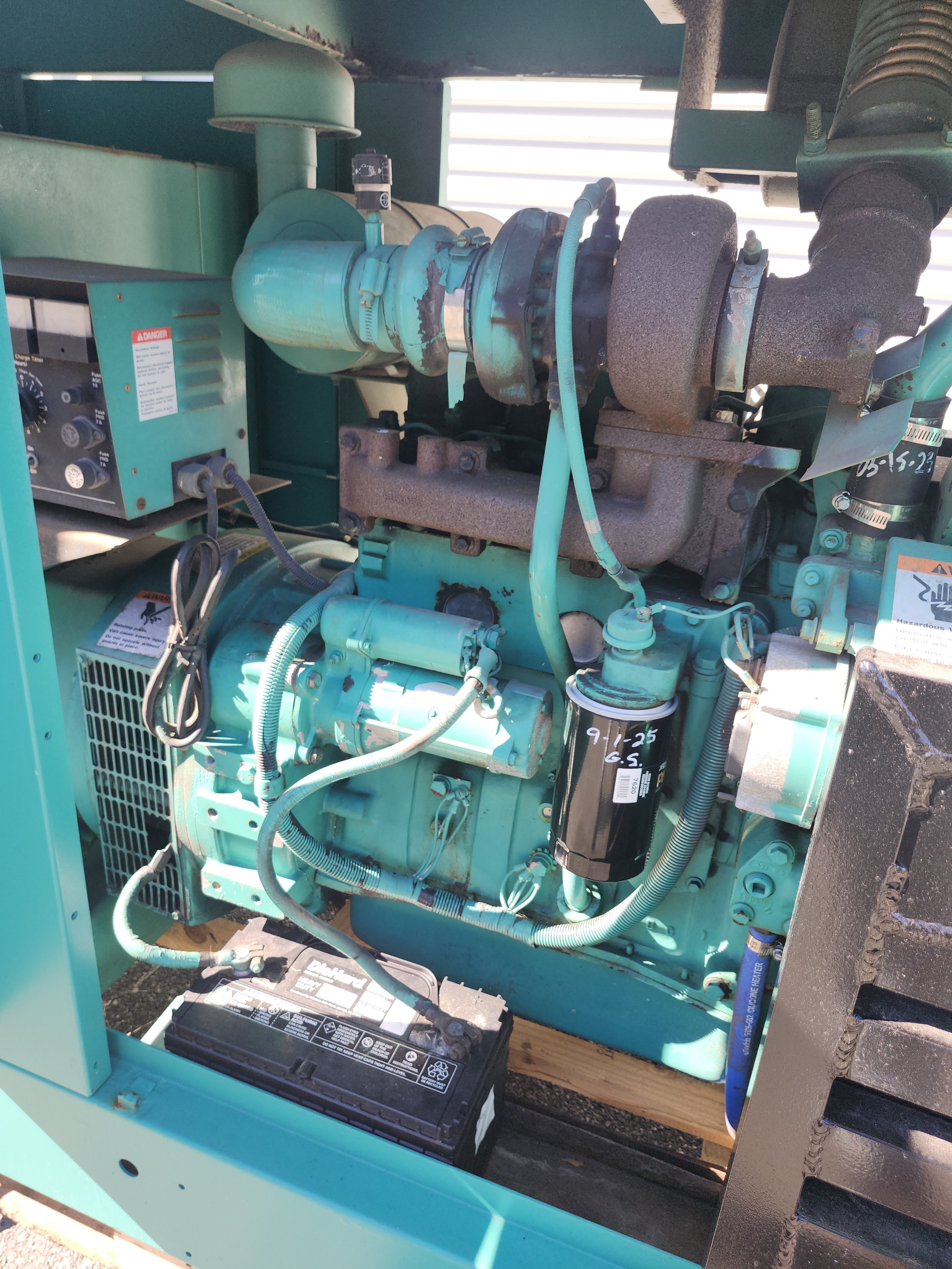 Close-up photo of a teal-colored industrial generator engine with various hoses, filters, and a battery visible.