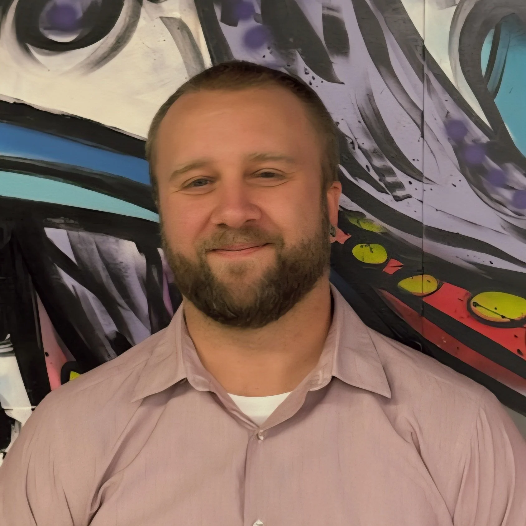 A man with a beard smiling, wearing a light pink button-up shirt, standing in front of colorful abstract mural art.