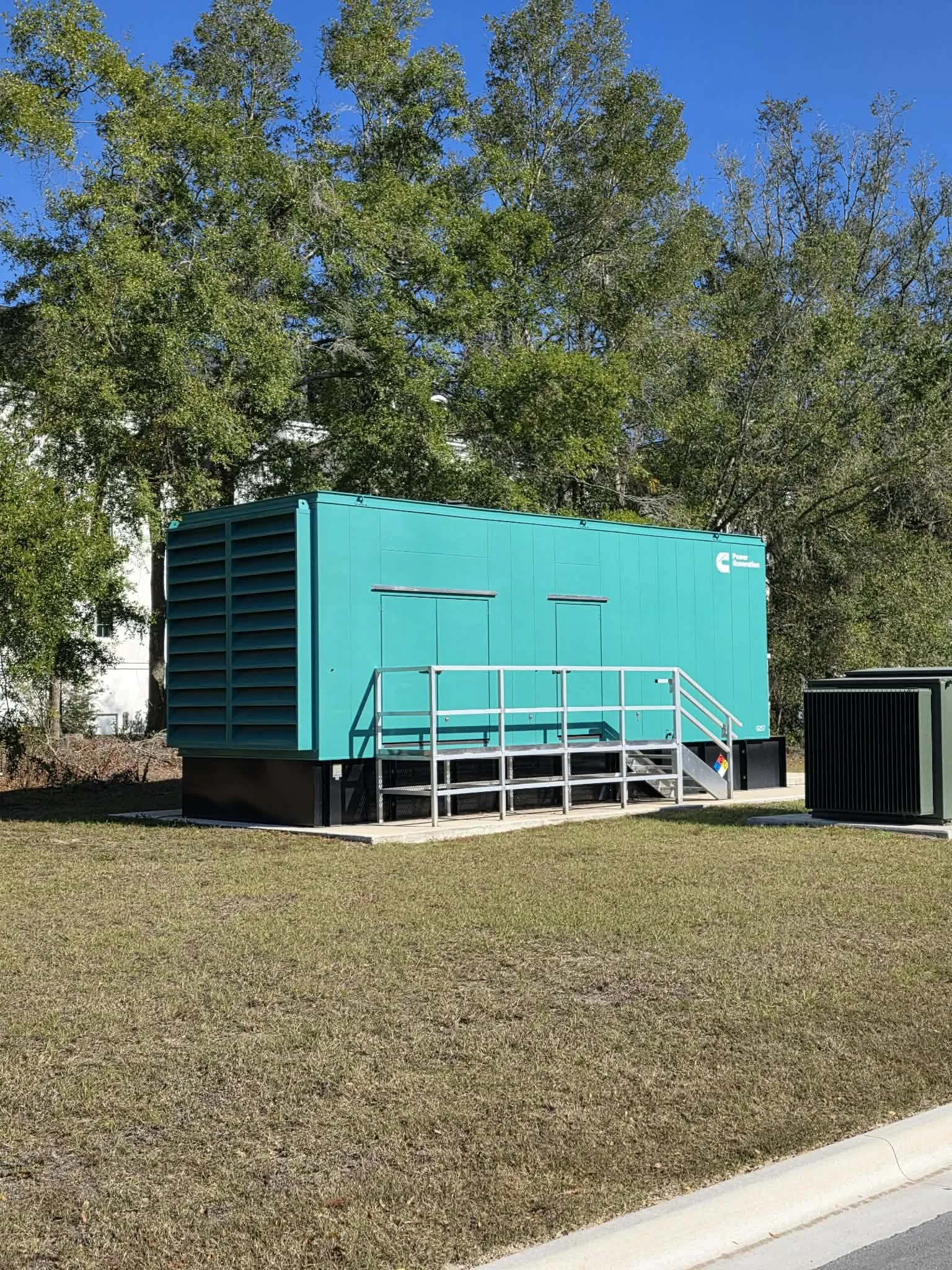 A large turquoise generator with a metal staircase in front, situated on a grassy area near a sidewalk and trees.