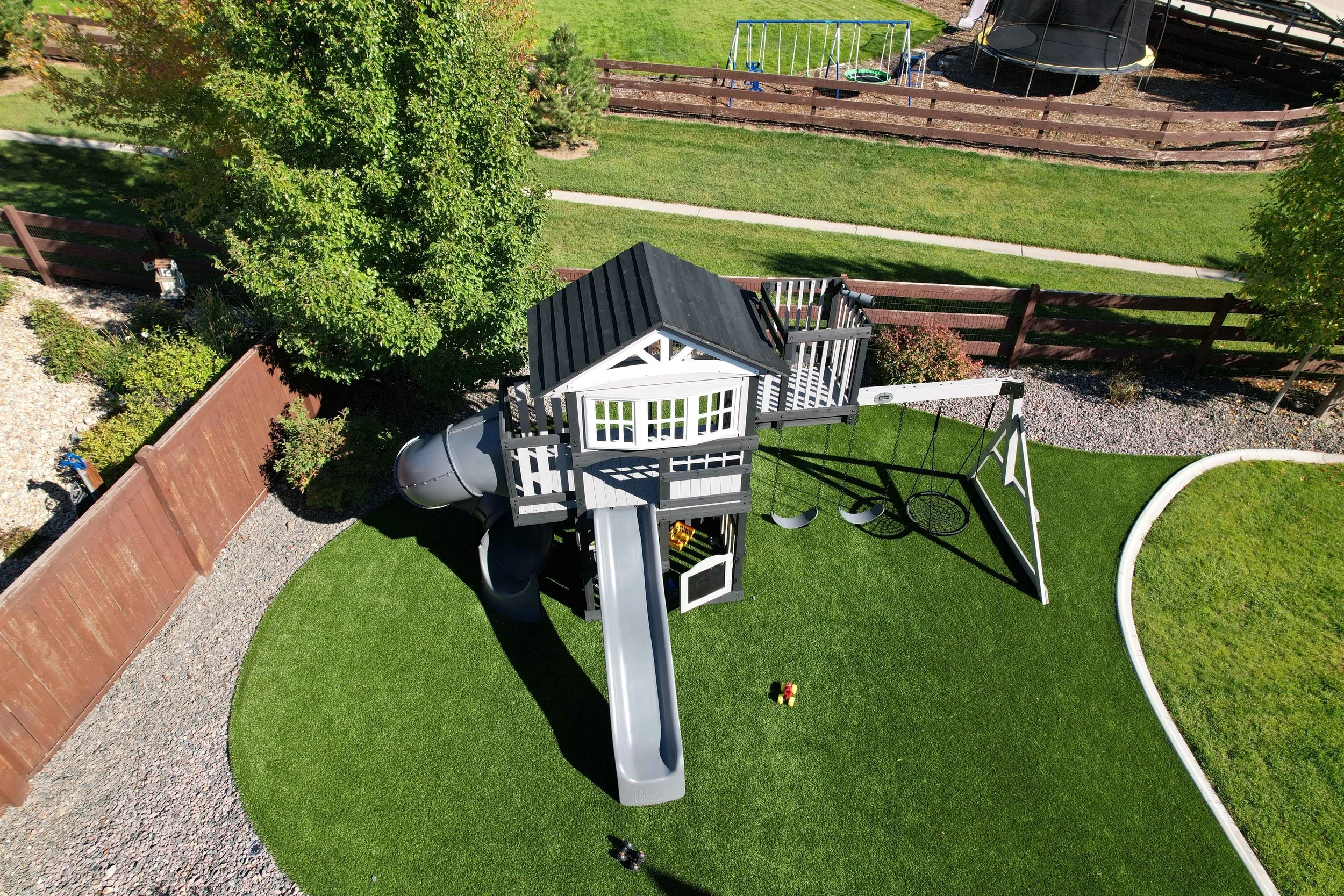 A backyard playground with a playhouse, slides, swings, and a trampoline, surrounded by green grass, trees, and a wooden fence.