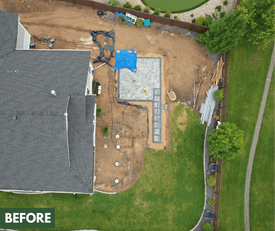A backyard with a house, soil, and a partially built paved patio or walkway. Construction materials and tools are visible, and the yard is fenced with green grass and trees.
