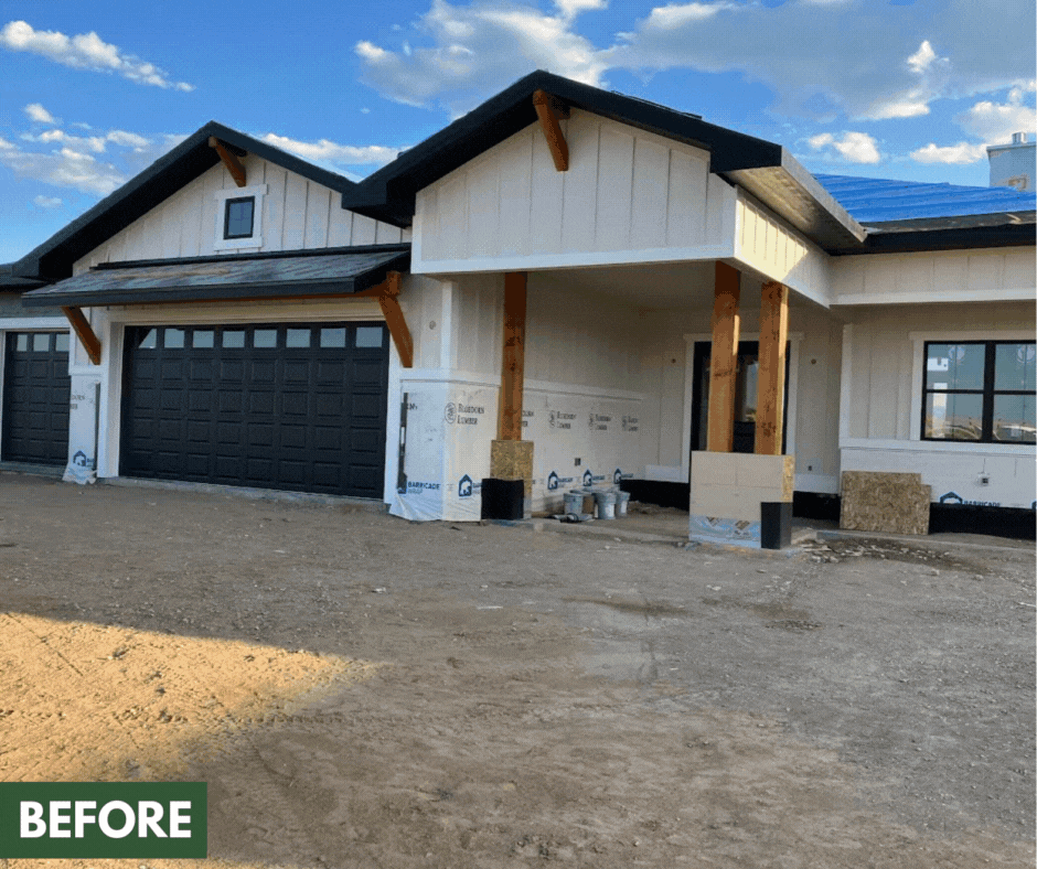 A house under construction with a two-car garage, white exterior siding, and wooden support beams in the front. The ground is bare dirt, and the sky is partly cloudy.