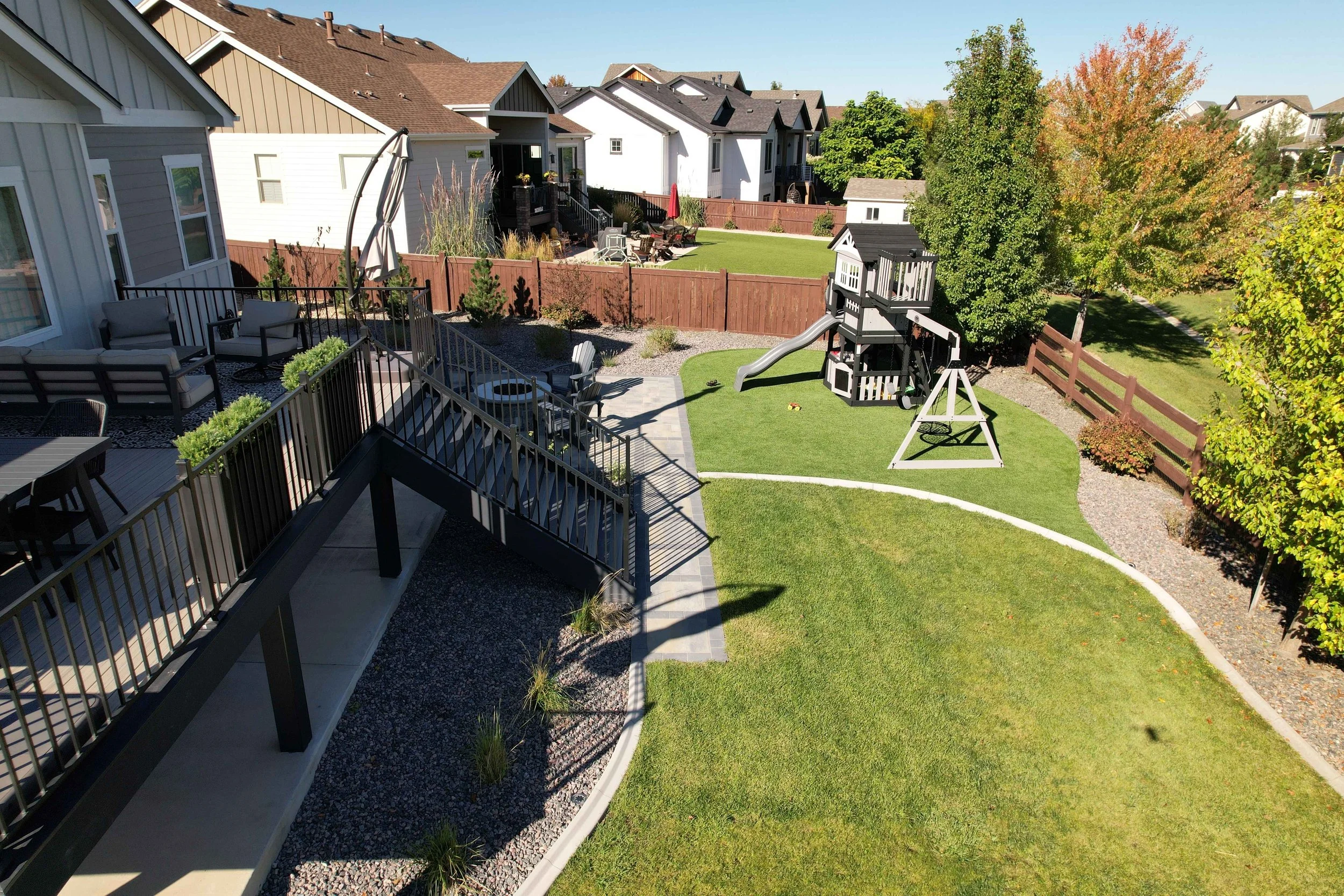 A backyard with a deck and patio furniture, a ramp leading to the grass, a playhouse with slide and swing set, trees, and neighboring houses in the background.