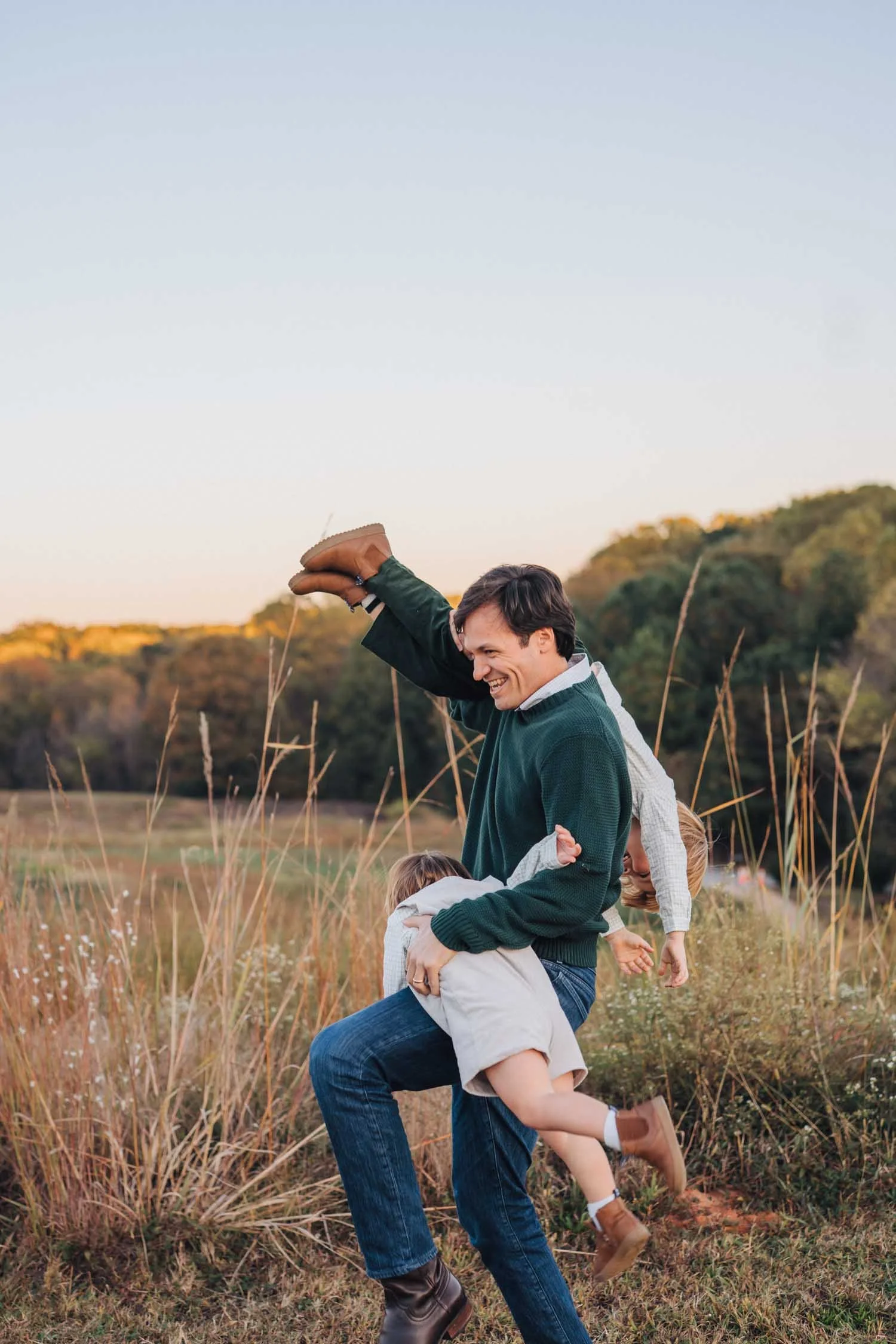 Father playing with two children and lifting them over his shoulders in a park during a playful family photography session in Raleigh, NC.