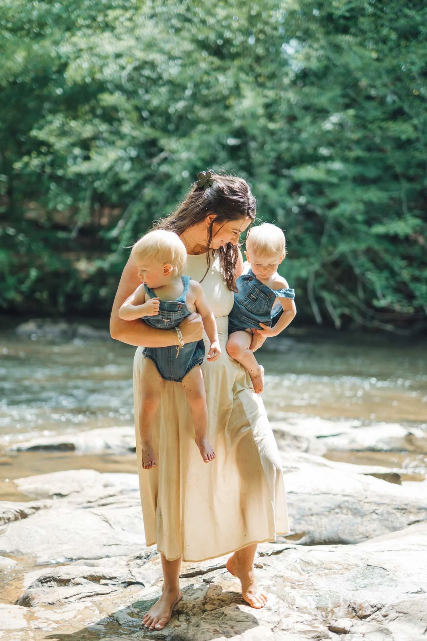 Mother walking through a forest creek holding two toddlers during a Locals family photography session in Raleigh, North Carolina.