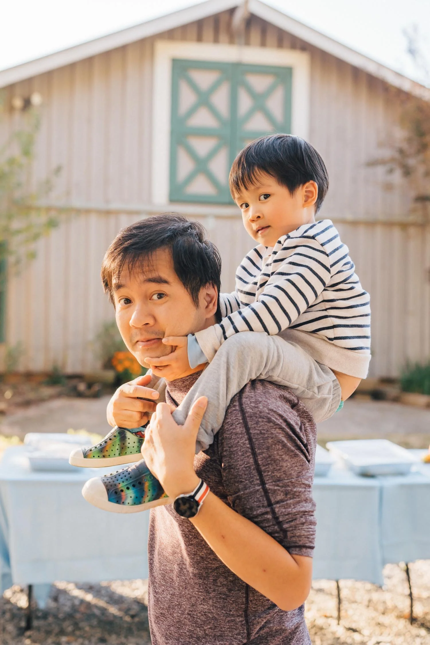 Father carrying his son on his shoulders outside near a barn during a lifestyle family photography session in North Carolina.