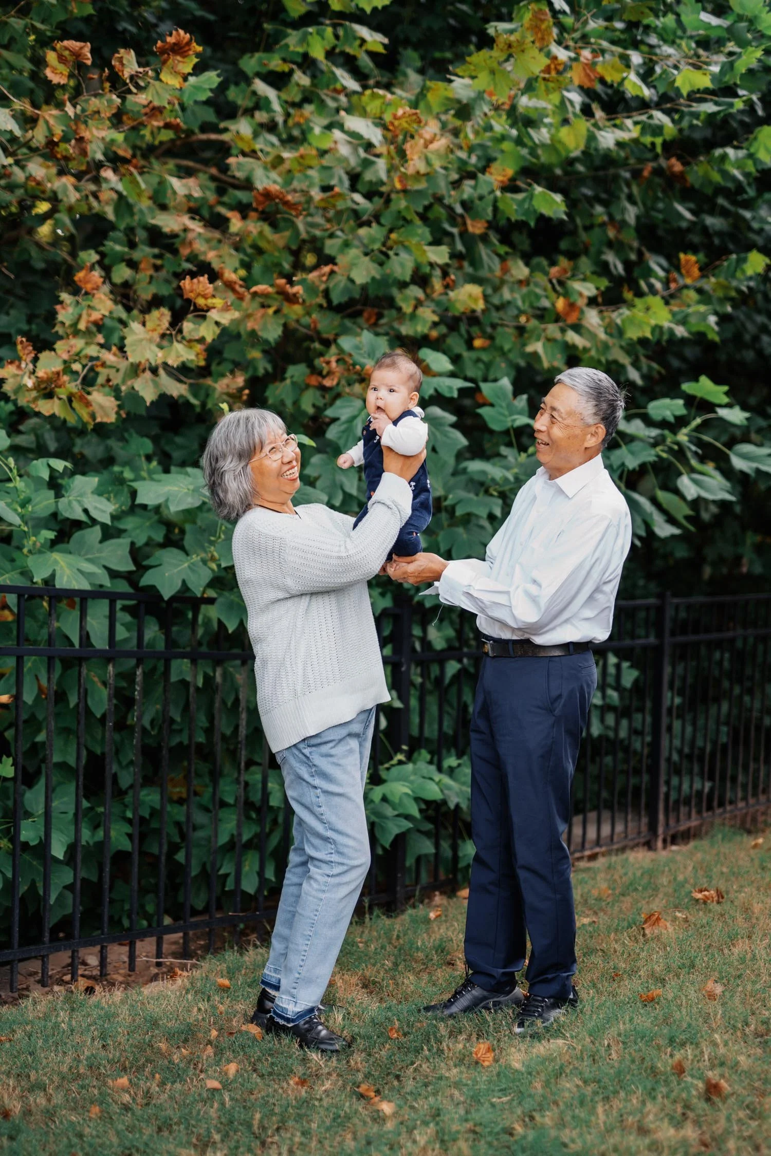 Two grandparents lifting a baby between them in a backyard during a multi-generational family photography session in Raleigh, NC.