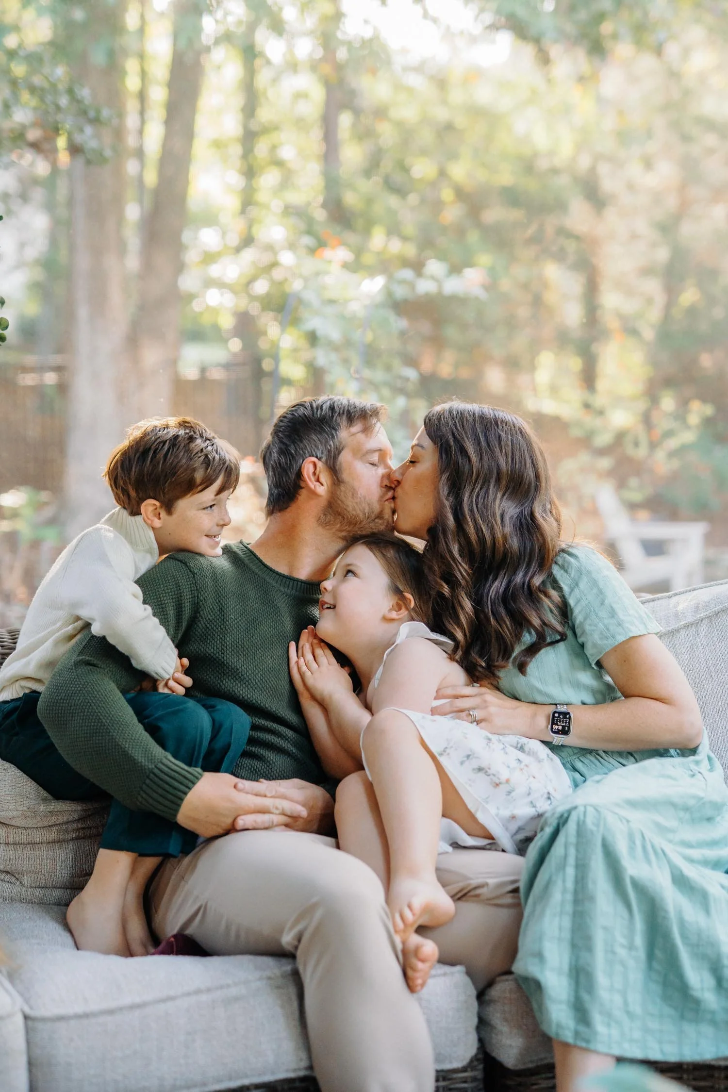 Parents kissing on the couch while their two children cuddle close during an in-home backyard family photography session in Raleigh, NC.