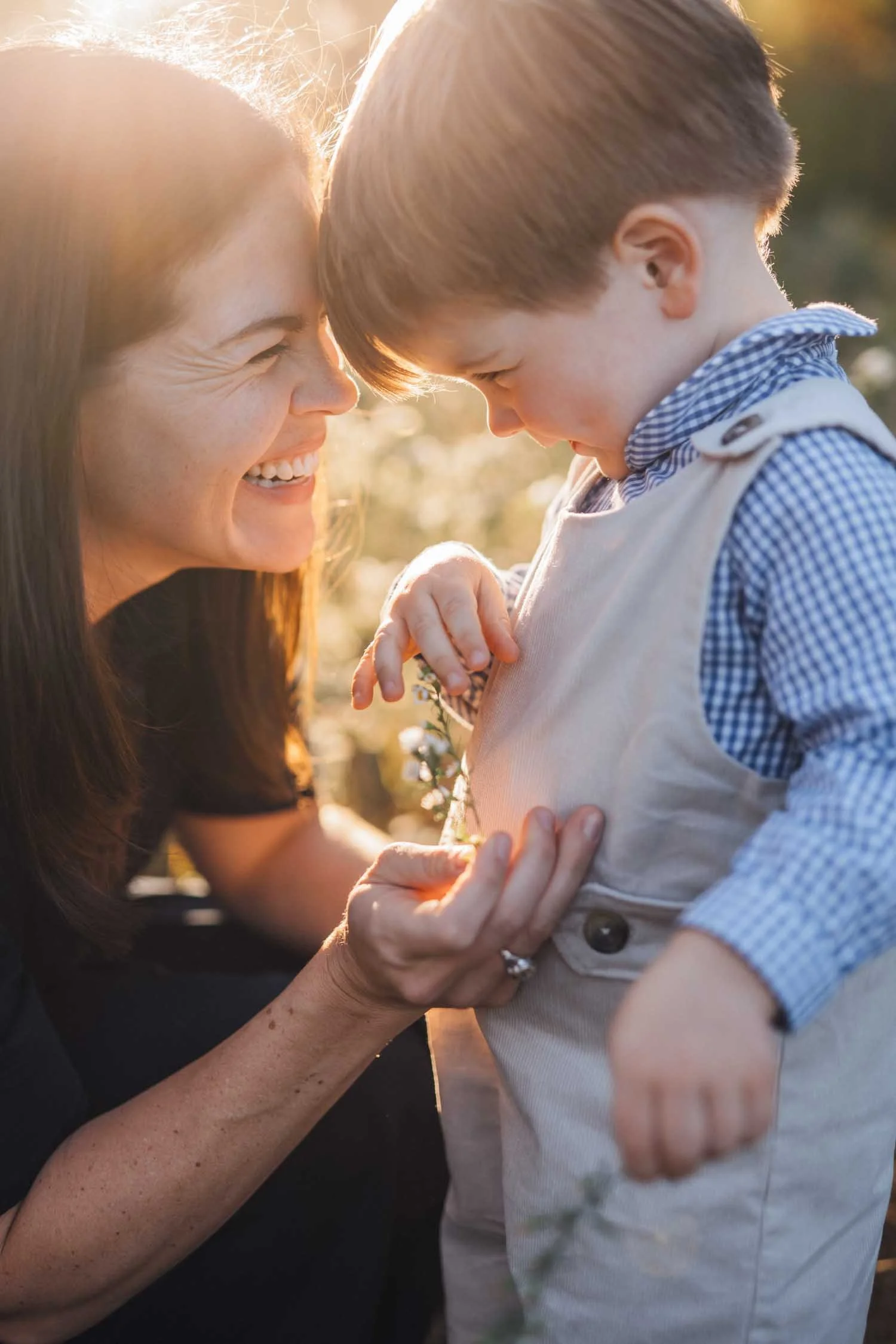 Mother and toddler smiling and laughing together while looking at a flower outdoors during a park family photography session in Raleigh–Durham.