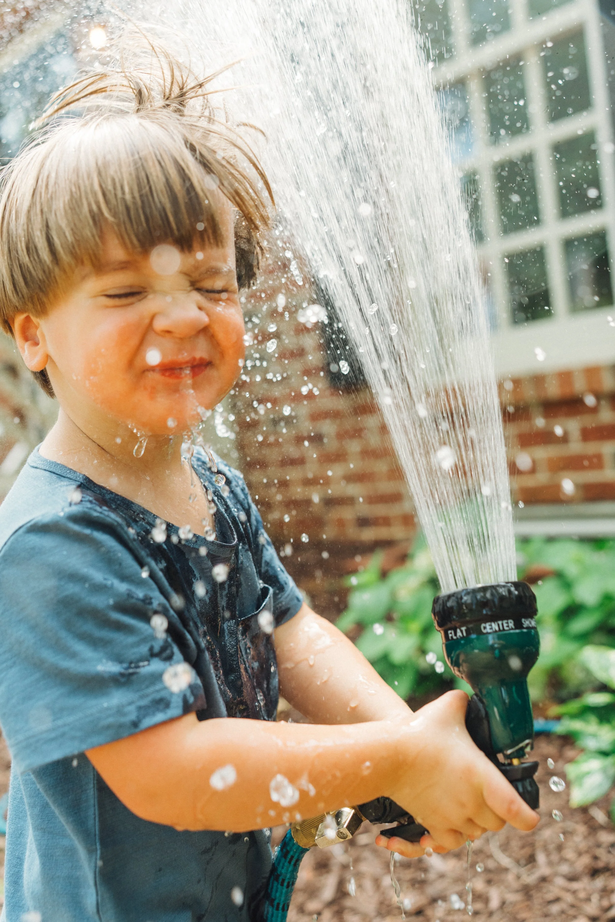 two year old boy in front yard spraying hose in his face joyfully during fun family photos in Raleigh, NC