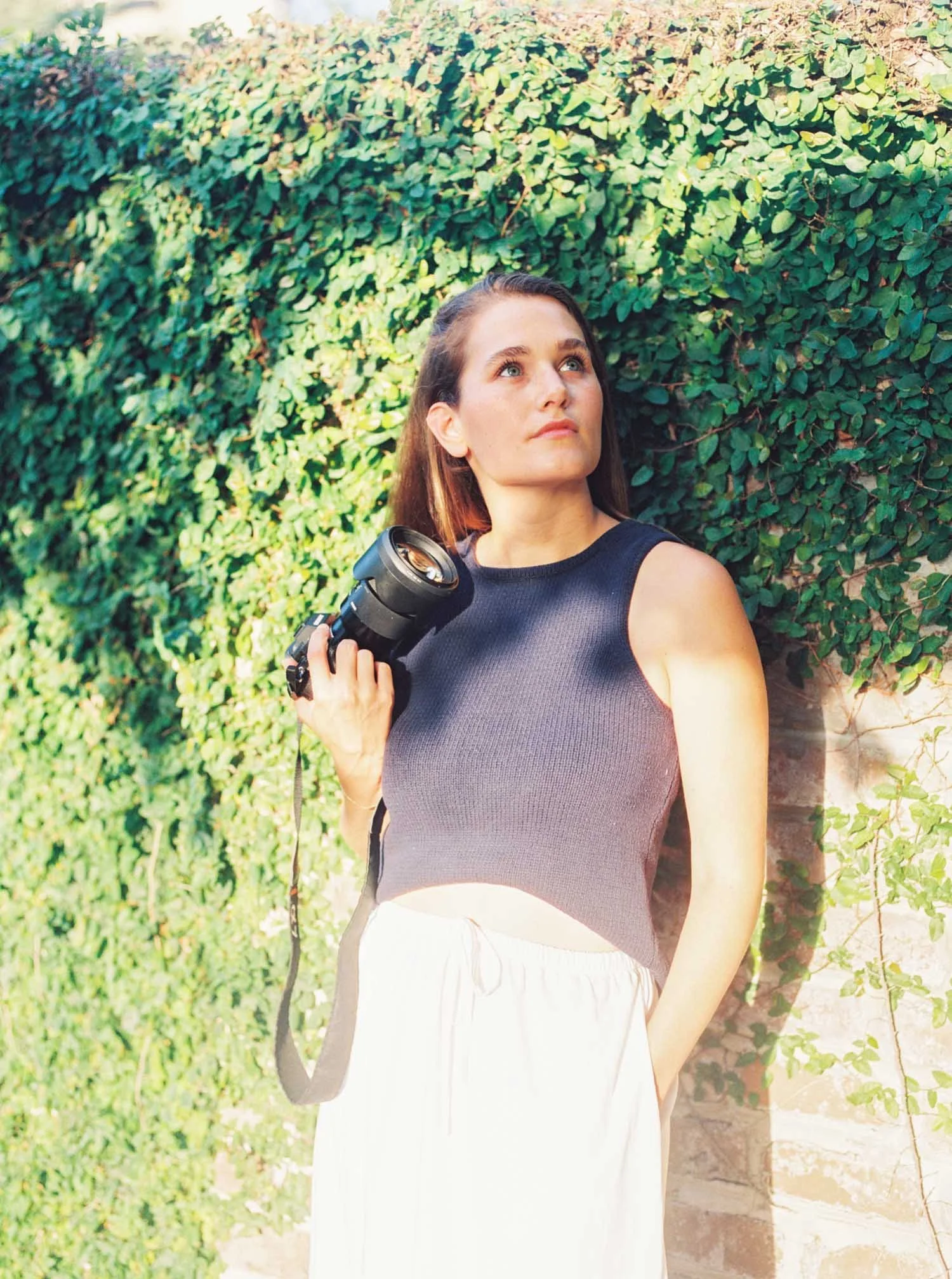 Autumn Brooke holding her camera and leaning against an ivy-covered wall outside, introducing the Raleigh, NC family photographer behind Autumn Brooke Photography.