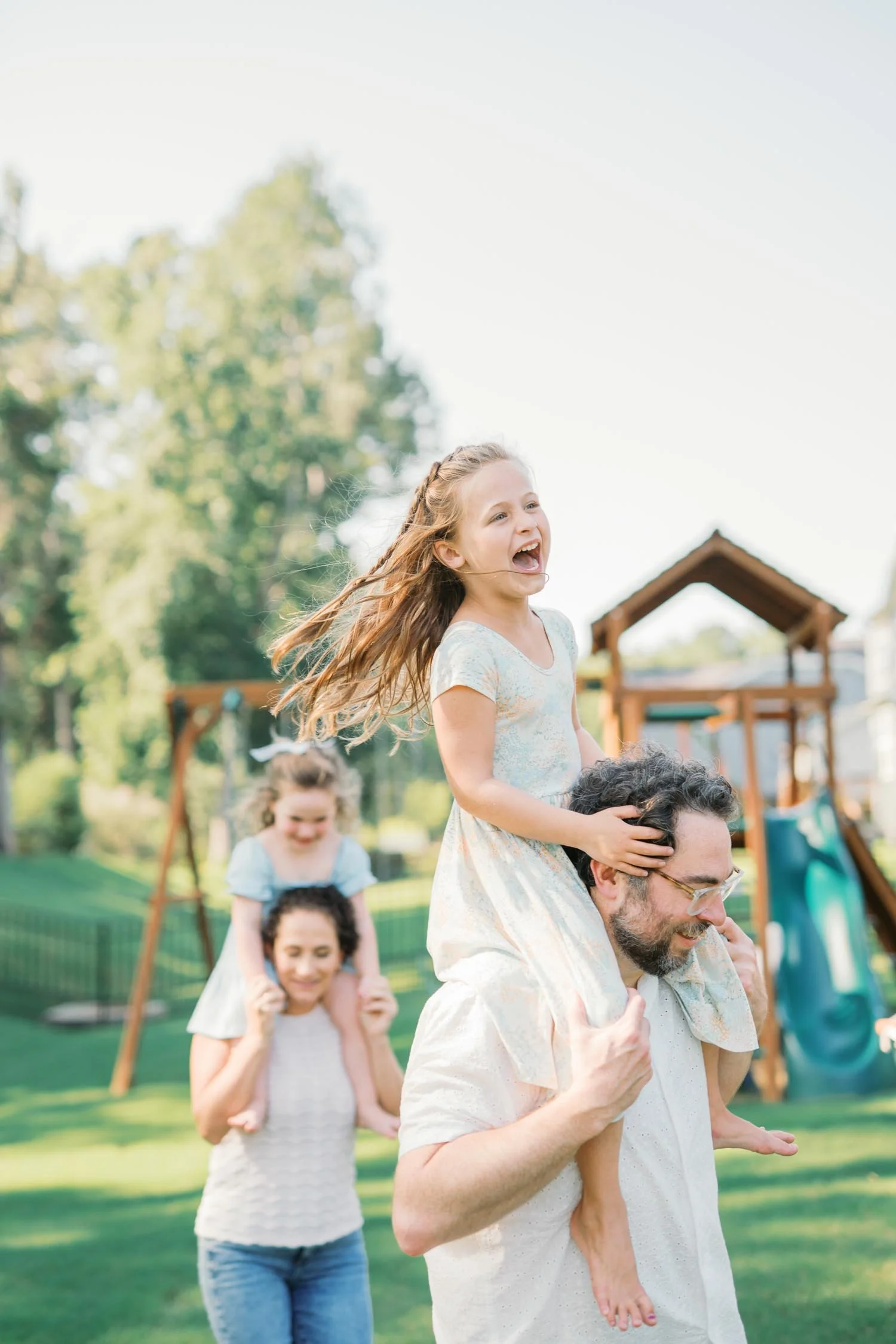 Two parents running through a playground with their children on their shoulders during a lifestyle family photography session in Raleigh, NC.