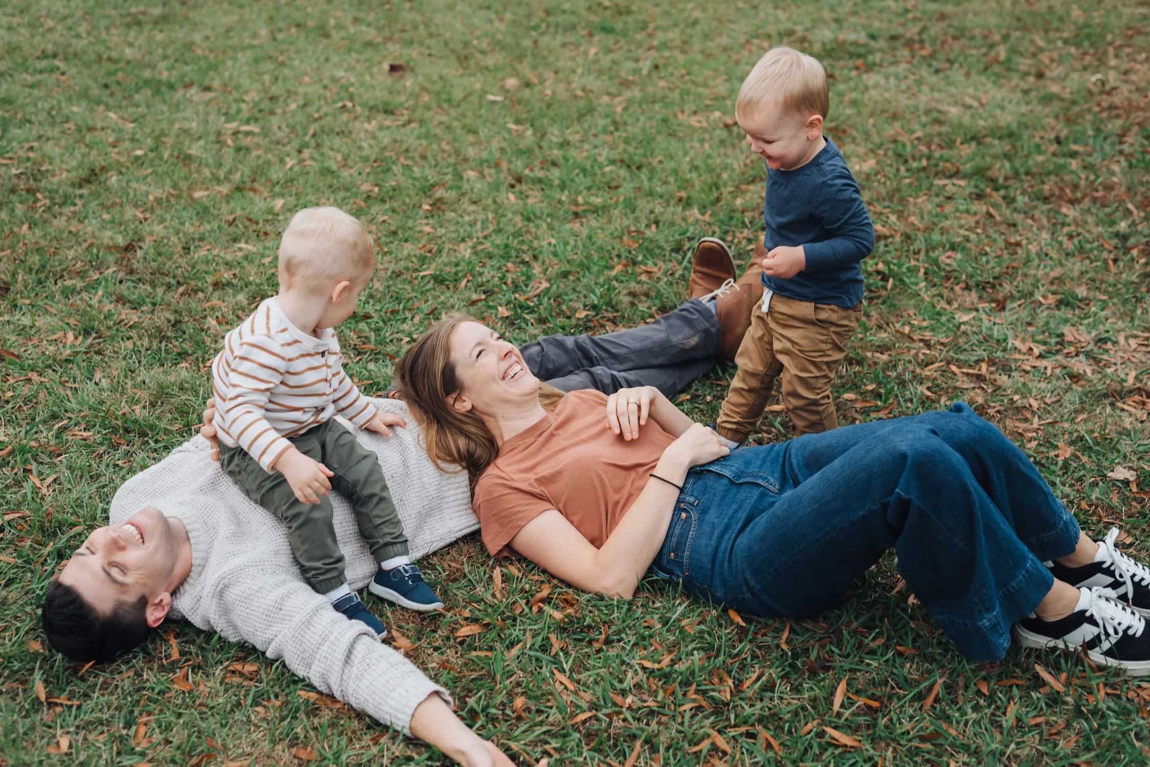 Mother and father lying in the grass while their two boys climb and play on them during a relaxed park family photography session.