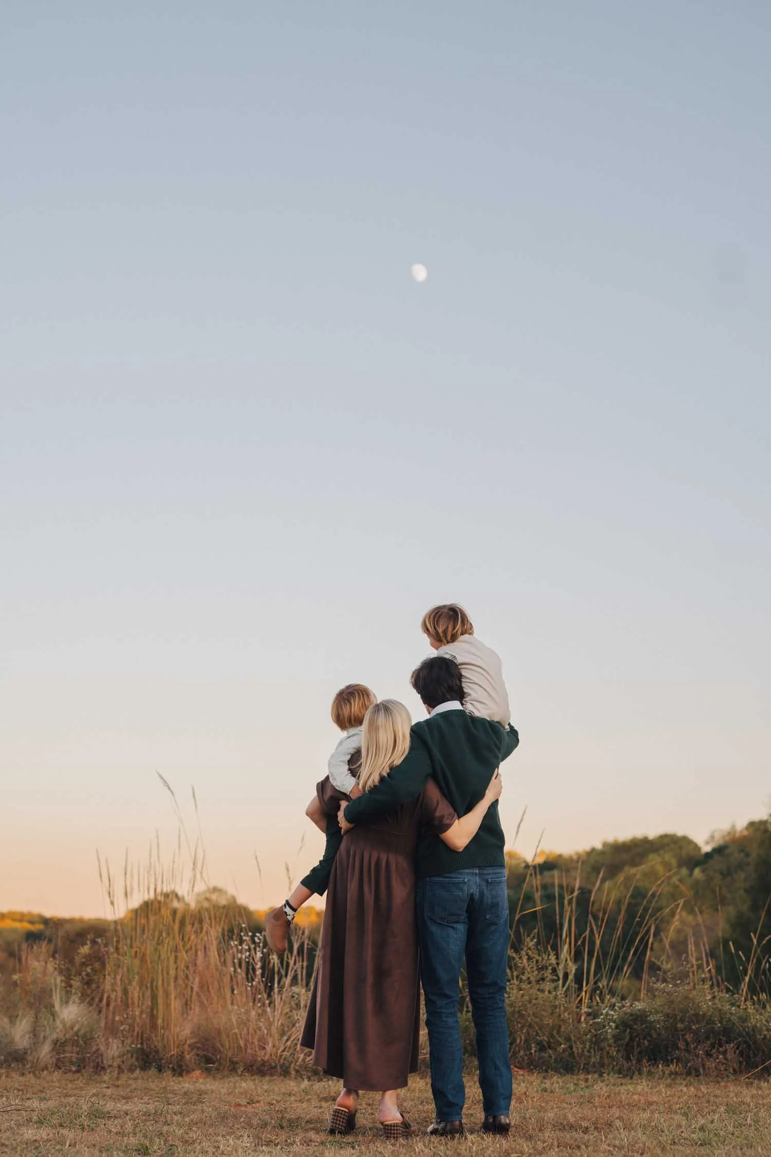Two parents lifting their children up together outdoors with the moon visible in the distance during a family photography session in Raleigh, North Carolina.