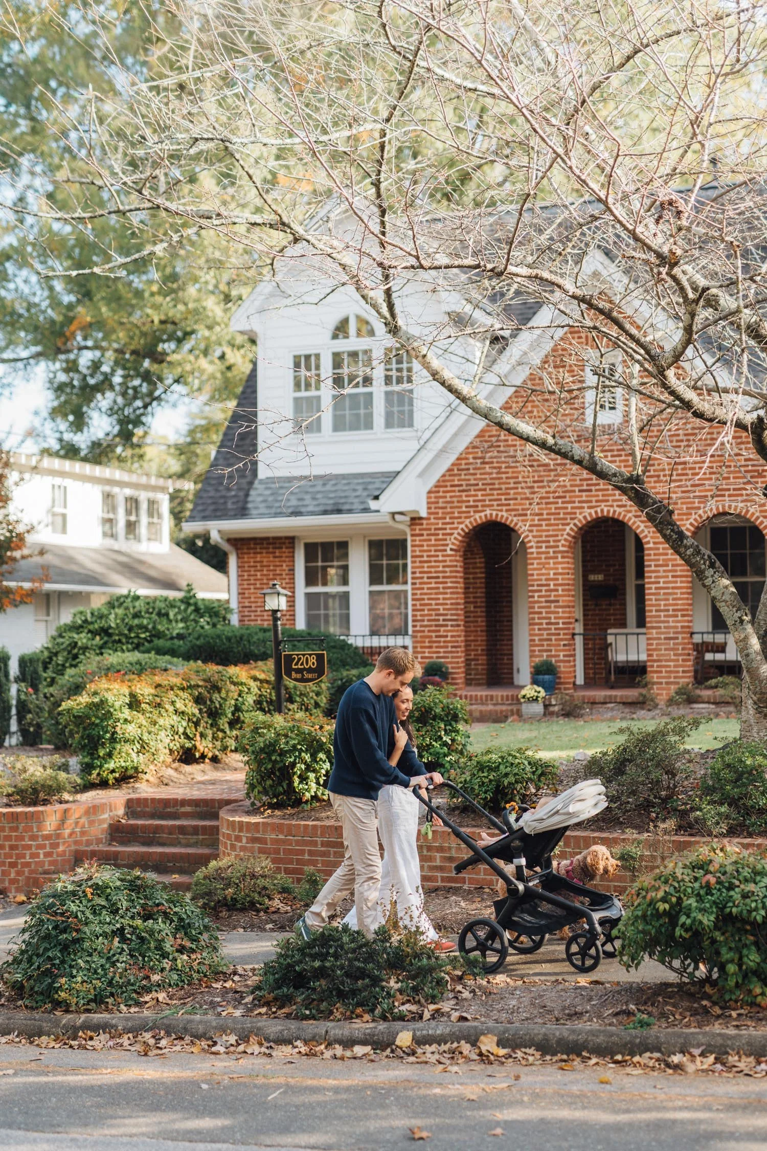 Parents walking their dog and pushing a stroller in front of their home during a family photography session by Autumn Brooke Photography in Raleigh, North Carolina.