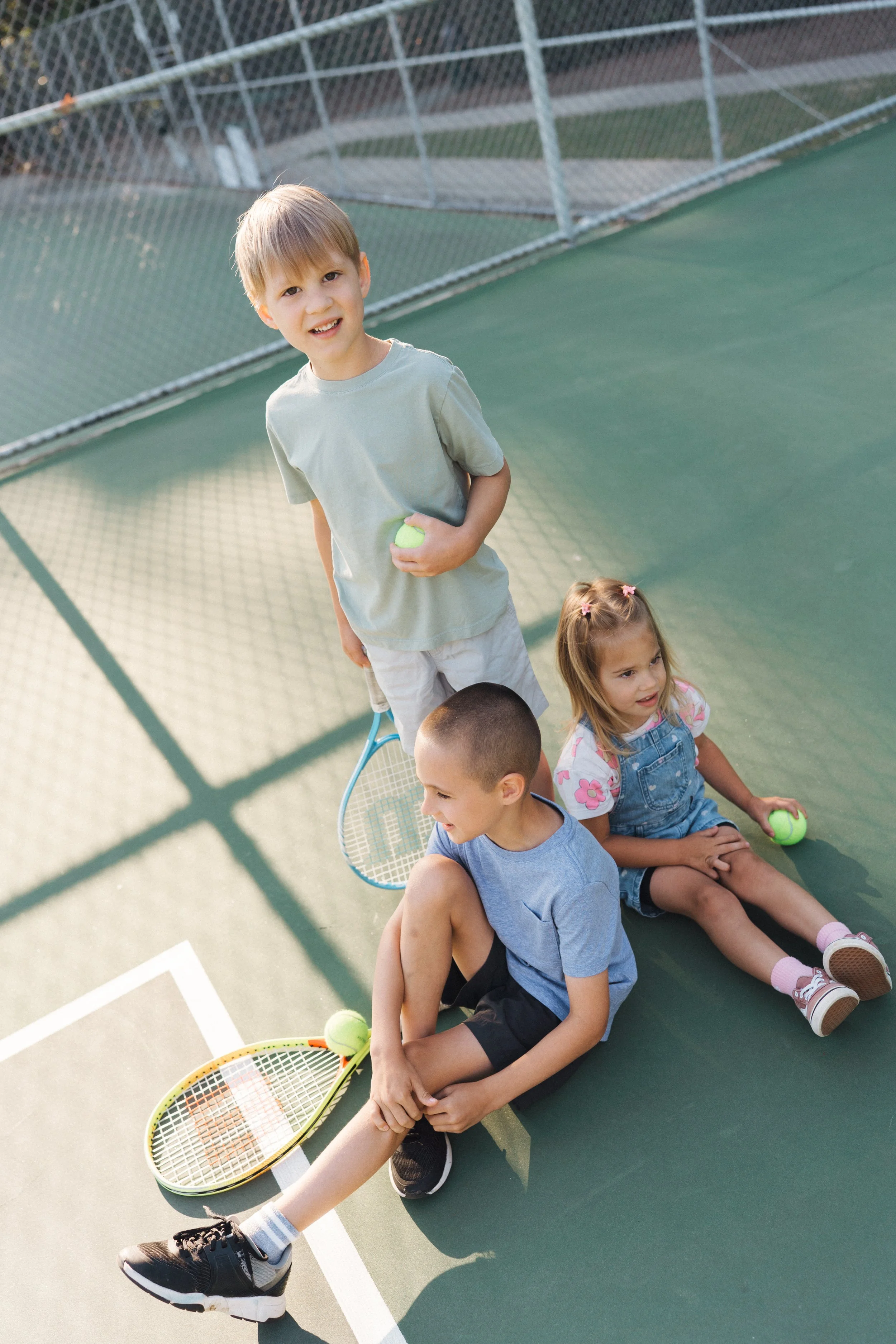 Three Siblings at a Tennis Court Holding Tennis Rackets and Smiling for Family Photography in Raleigh, NC