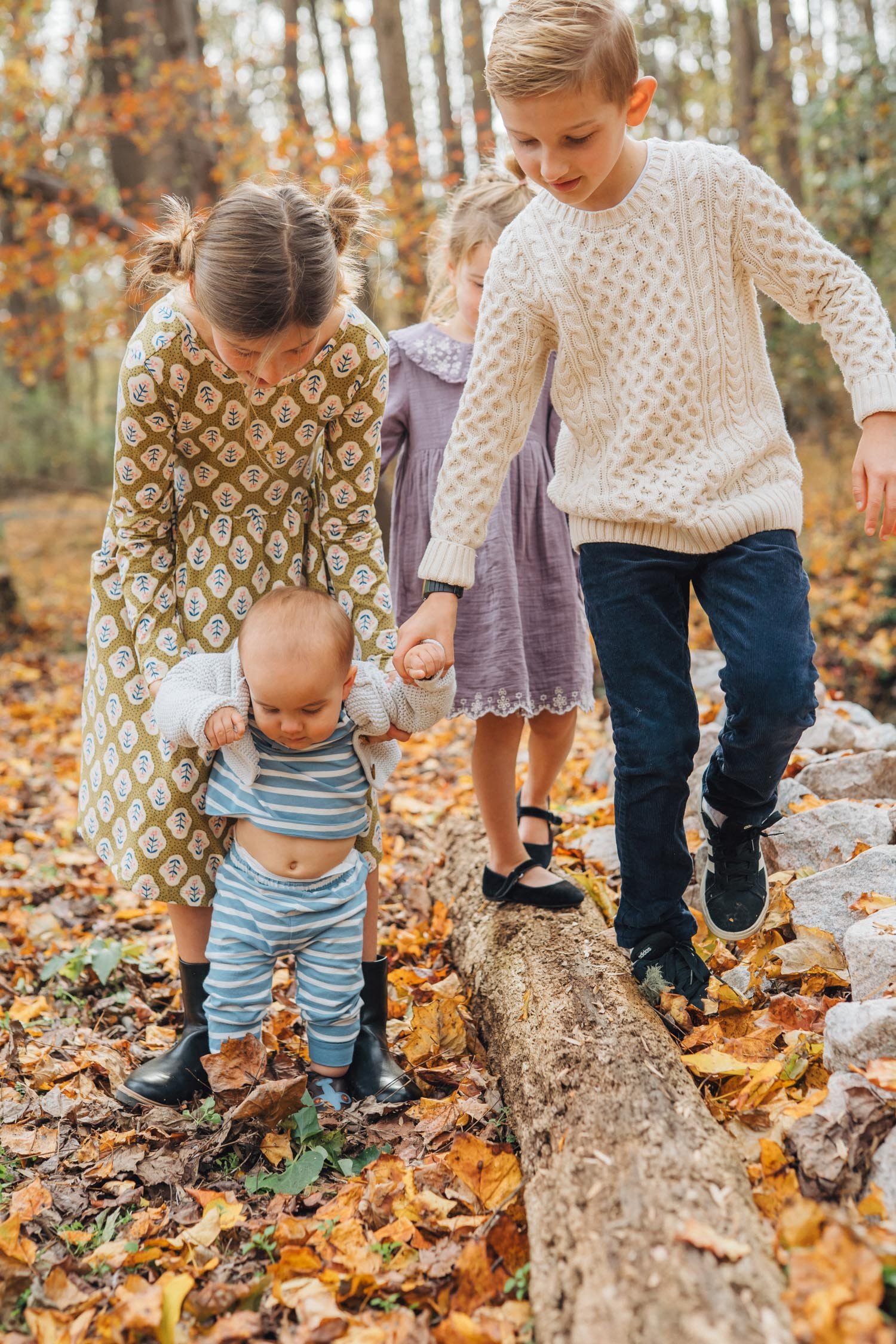 Three children holding hands with a baby while walking through the woods during a backyard family photography session in Raleigh–Durham.