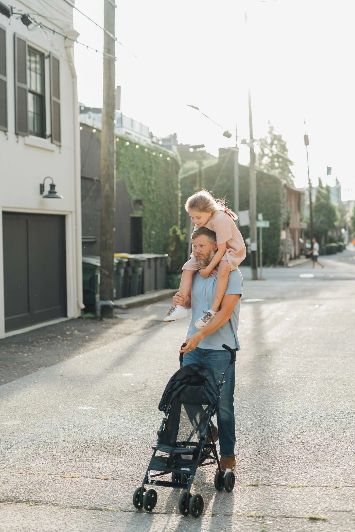 Father pushing a stroller while carrying a toddler on his shoulders in a walkable downtown area during a Locals family photography session in the Raleigh–Durham area.