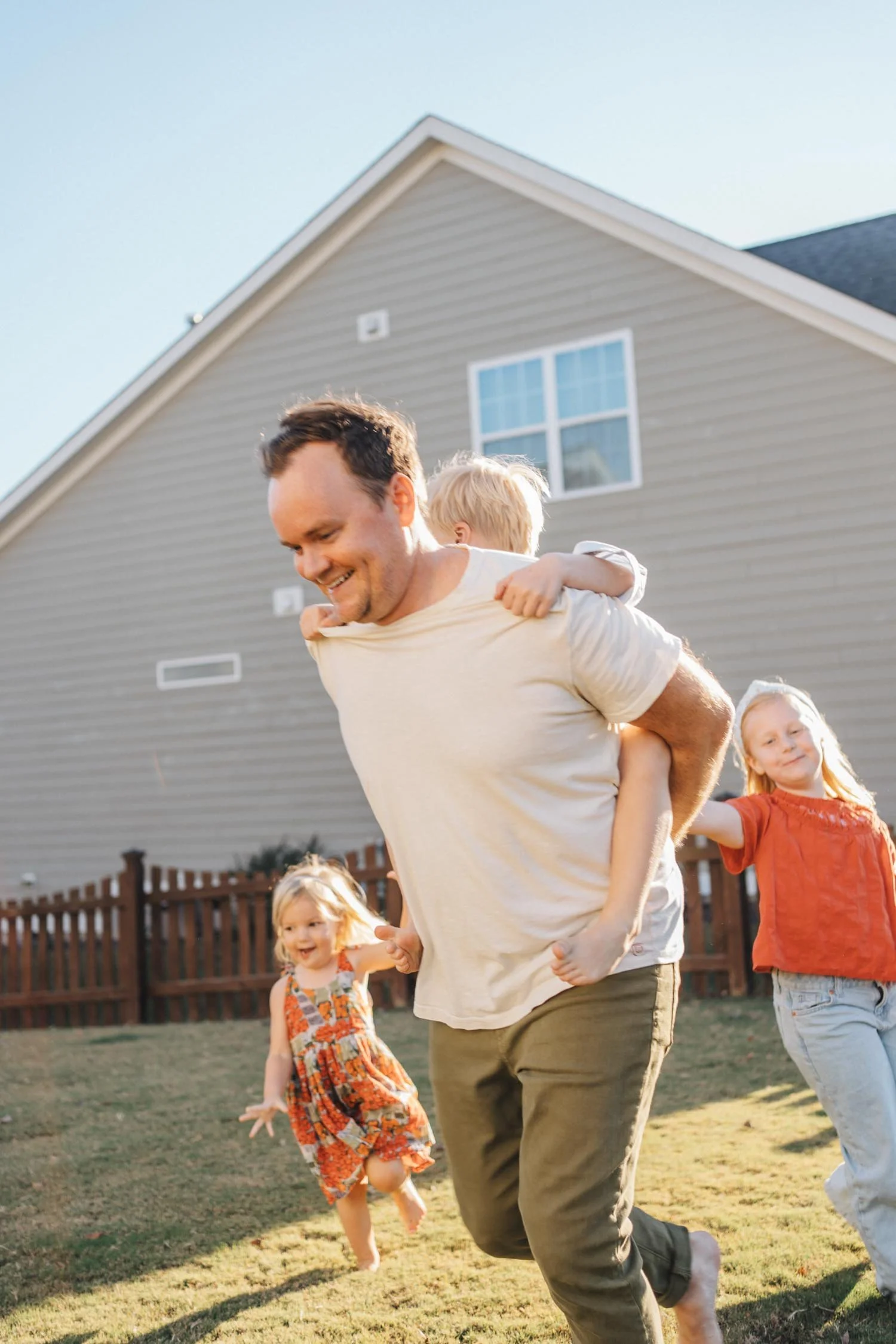 Father running through the backyard with one child on his back while two others run behind during a signature backyard family photography session in Raleigh, North Carolina.