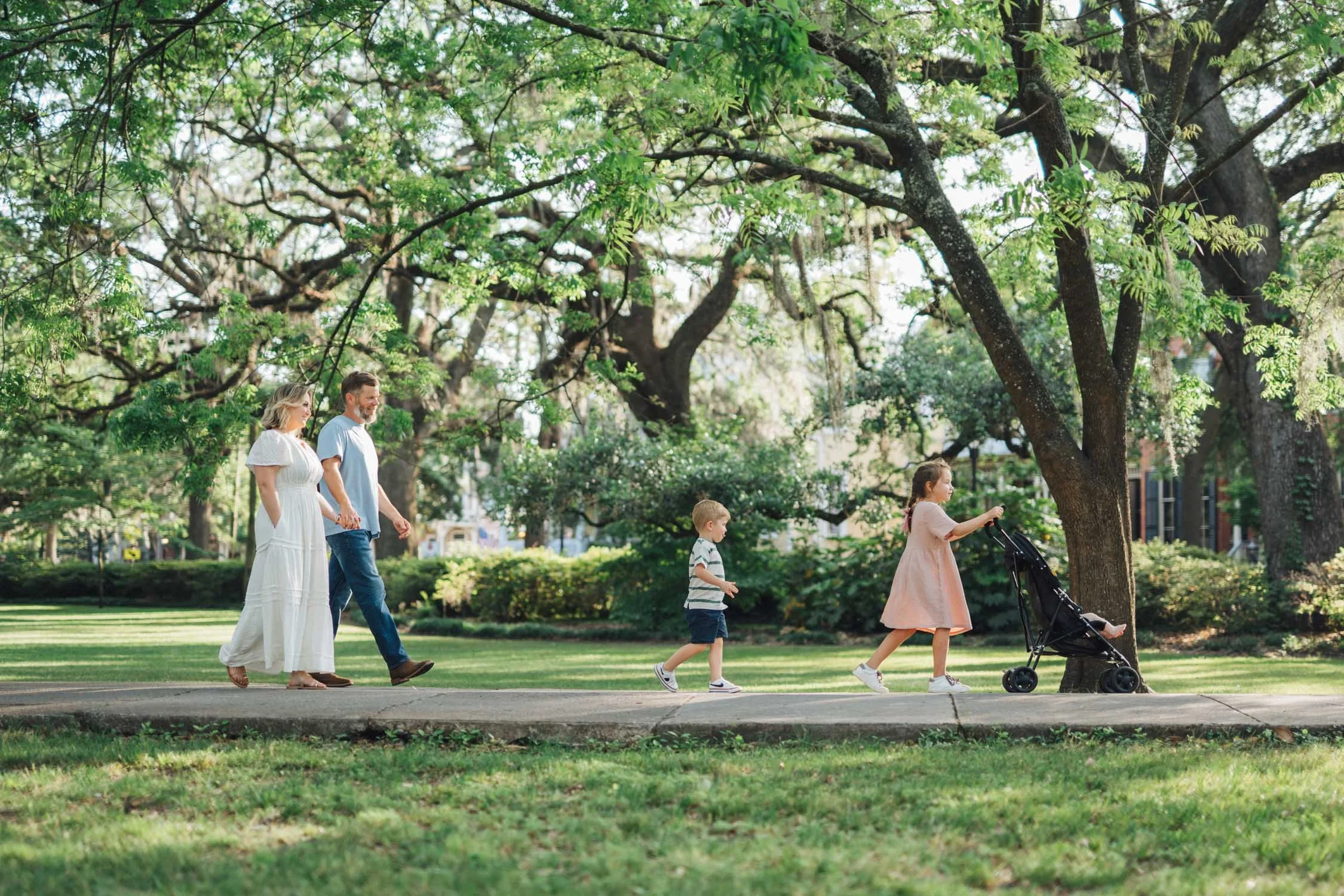Family of four walking together while pushing a stroller through a park during a lifestyle family photography session in Raleigh, NC.
