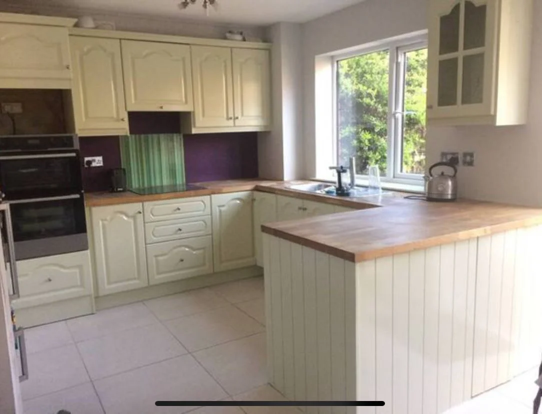 Kitchen with cream-colored cabinets, wooden countertops, and a window overlooking green trees. Contains a kettle, sink, and built-in oven.