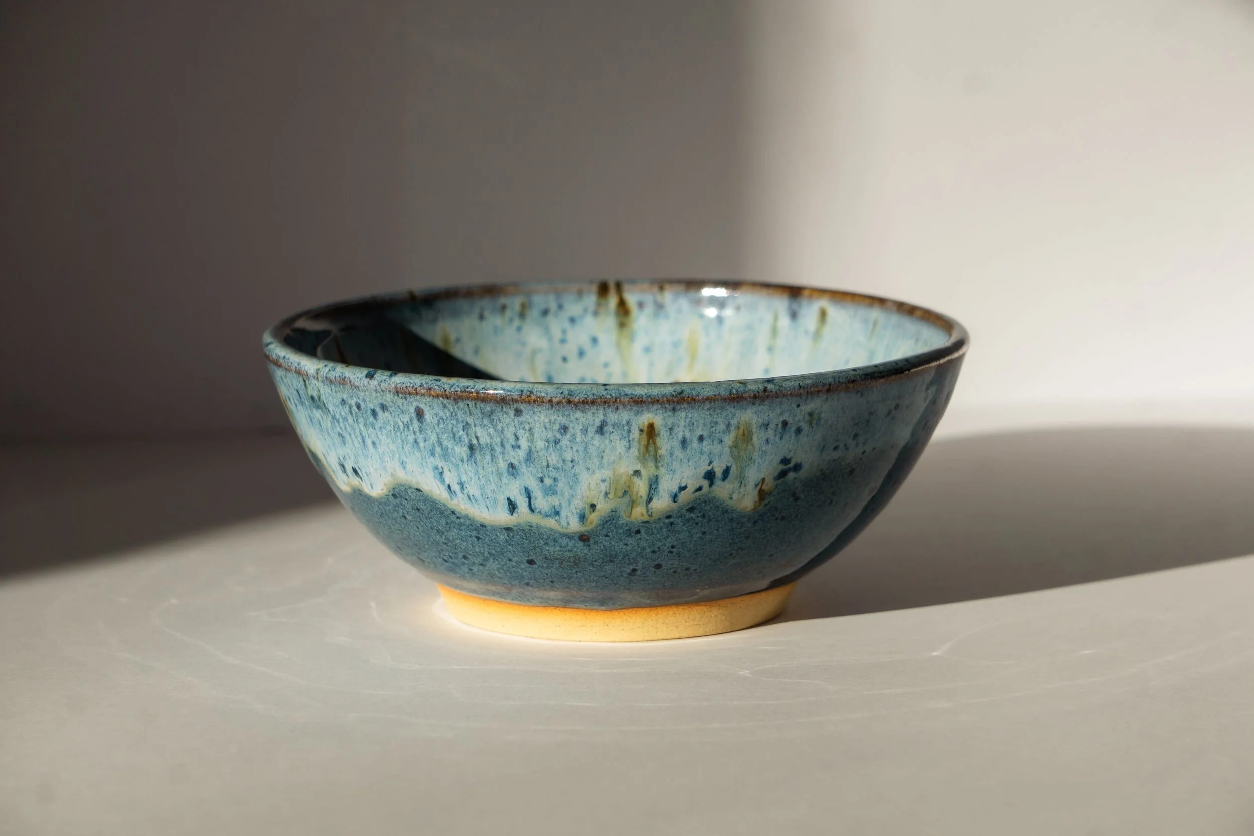 A ceramic bowl with a blue glaze and earthy tones, placed on a white surface with a soft shadow, illuminated by natural light.
