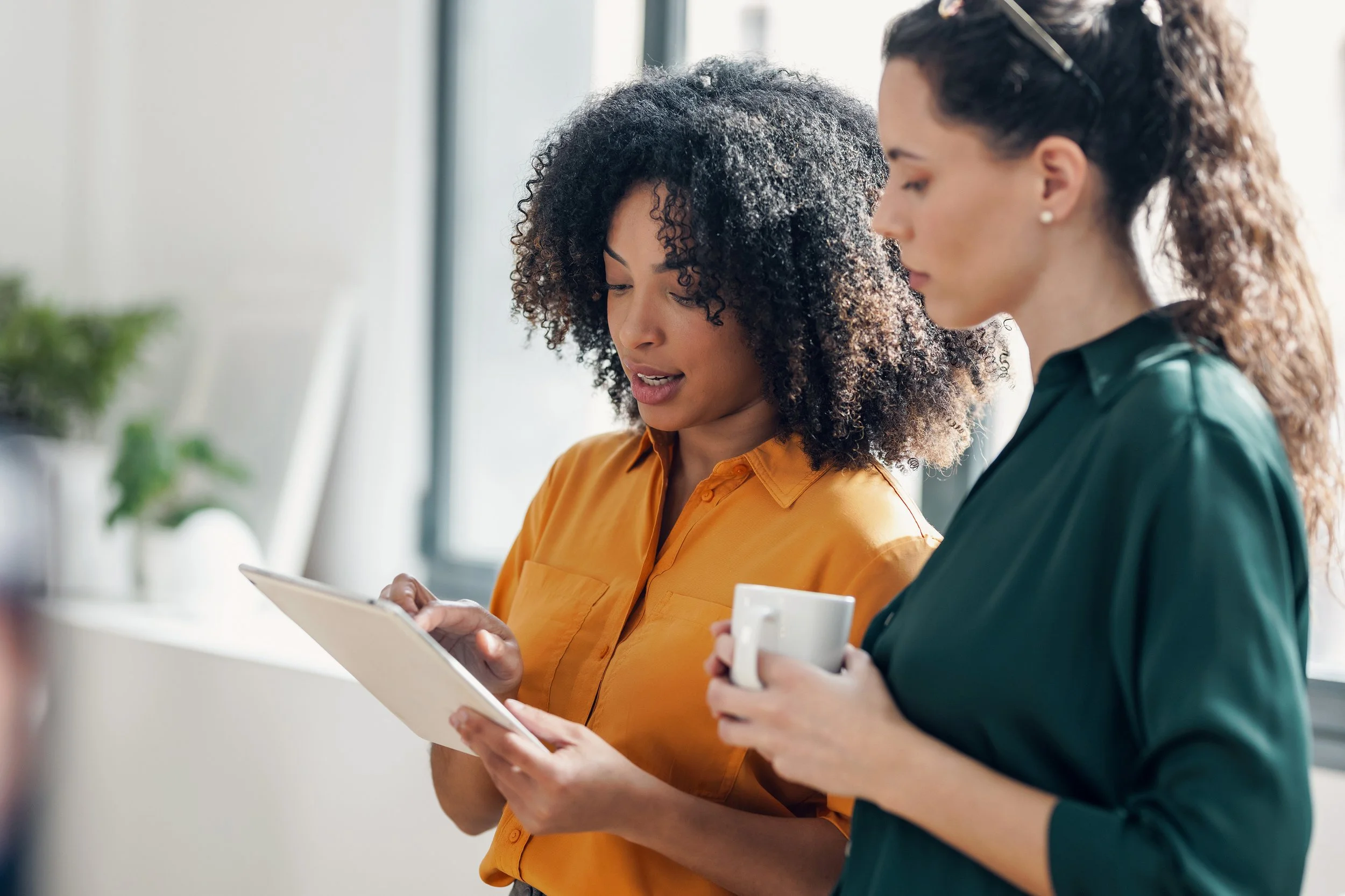 Two women standing together, one showing something on a tablet to the other while holding a coffee mug.