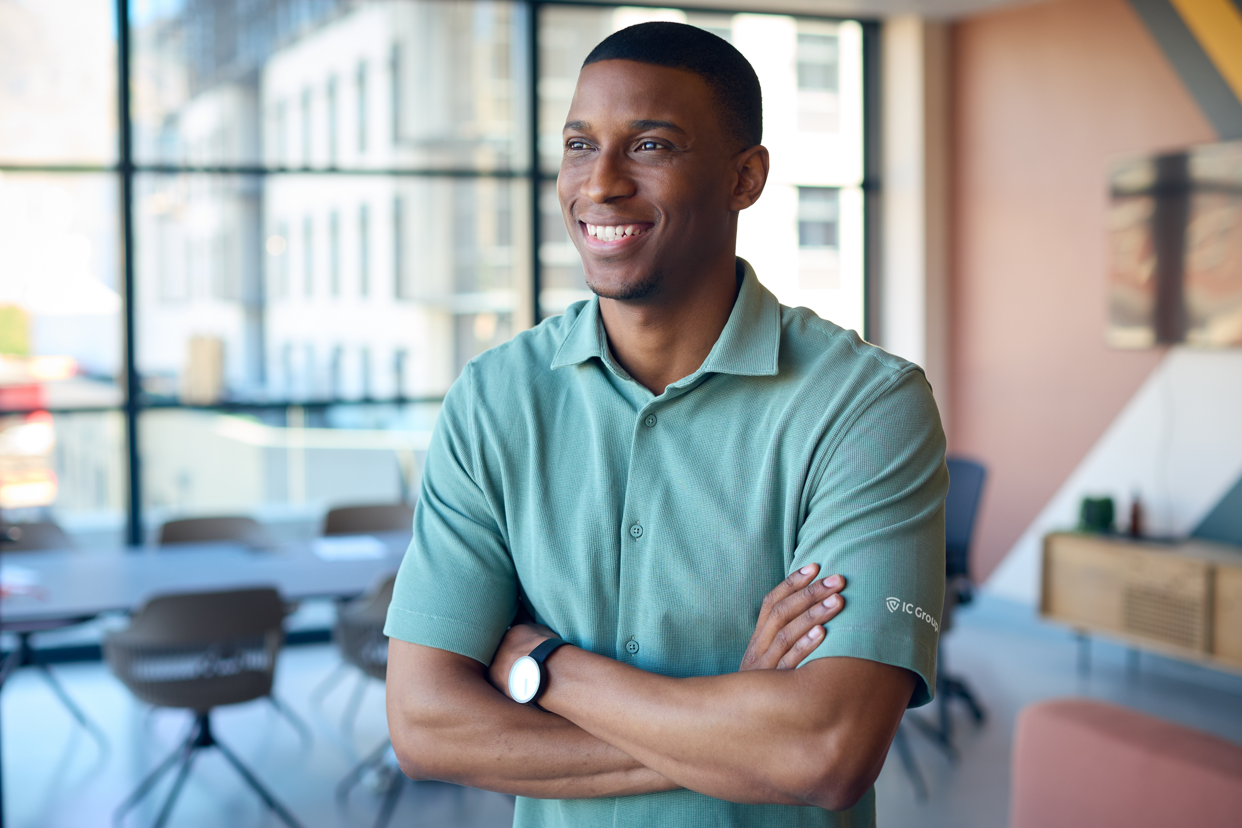A young man with dark skin and short haircut smiling, standing with arms crossed in a modern office space with large windows and a conference table.