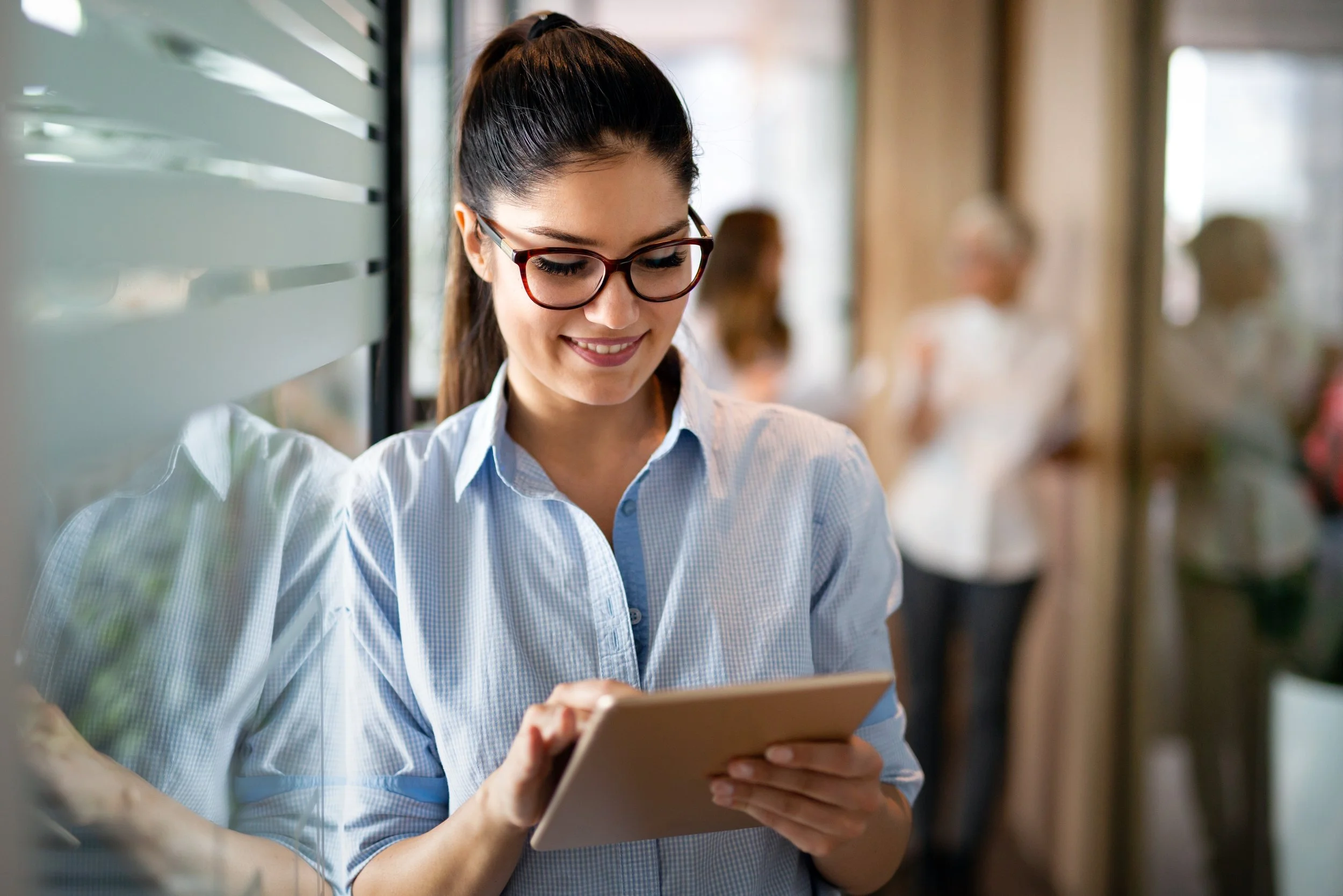 A young woman with dark hair tied back, wearing glasses and a light blue button-up shirt, smiling while looking at a tablet in a social setting or office.