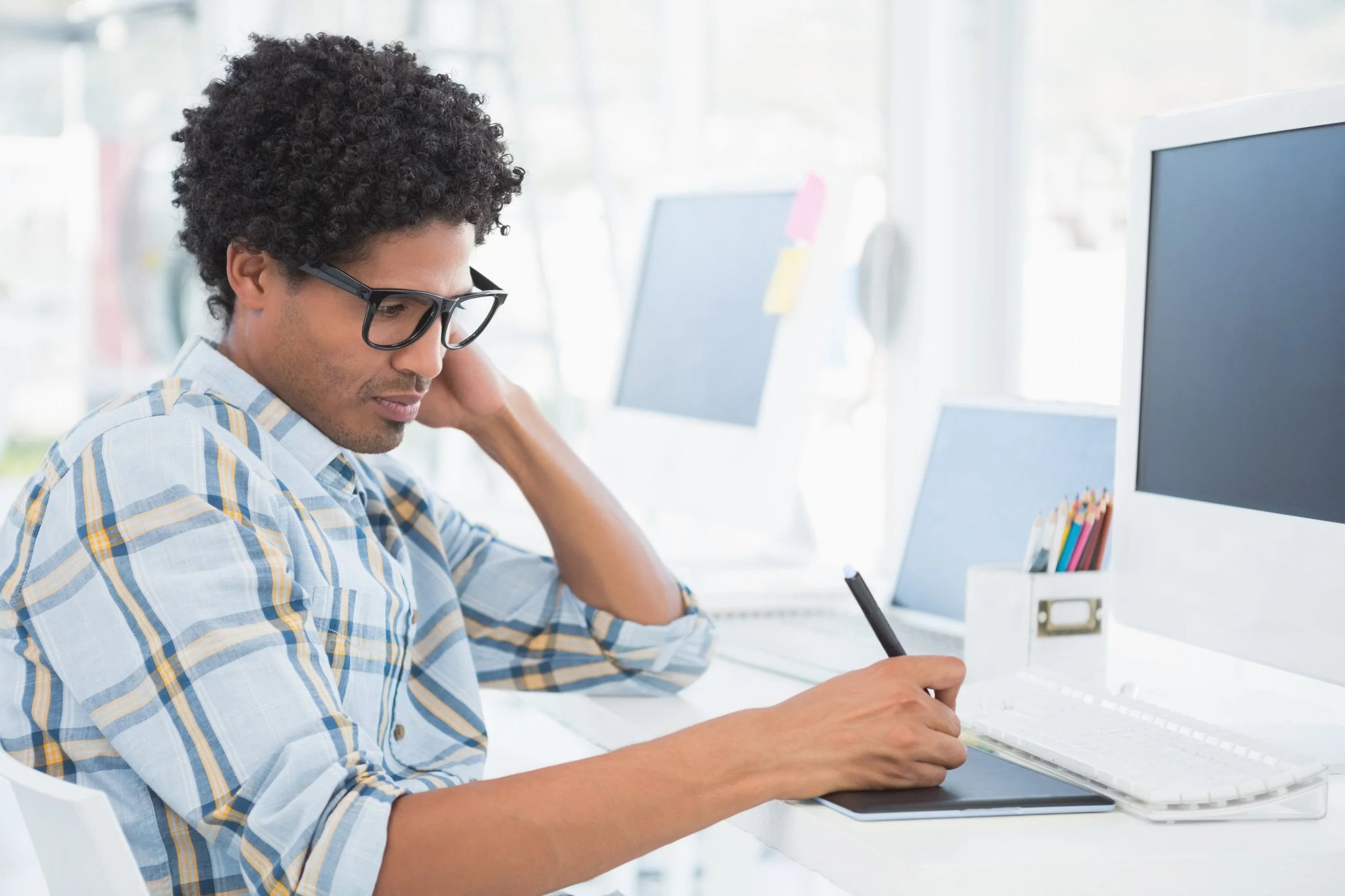 Man wearing glasses using a stylus on a graphics tablet while working at a desk with a computer monitors and office supplies in a bright modern office.