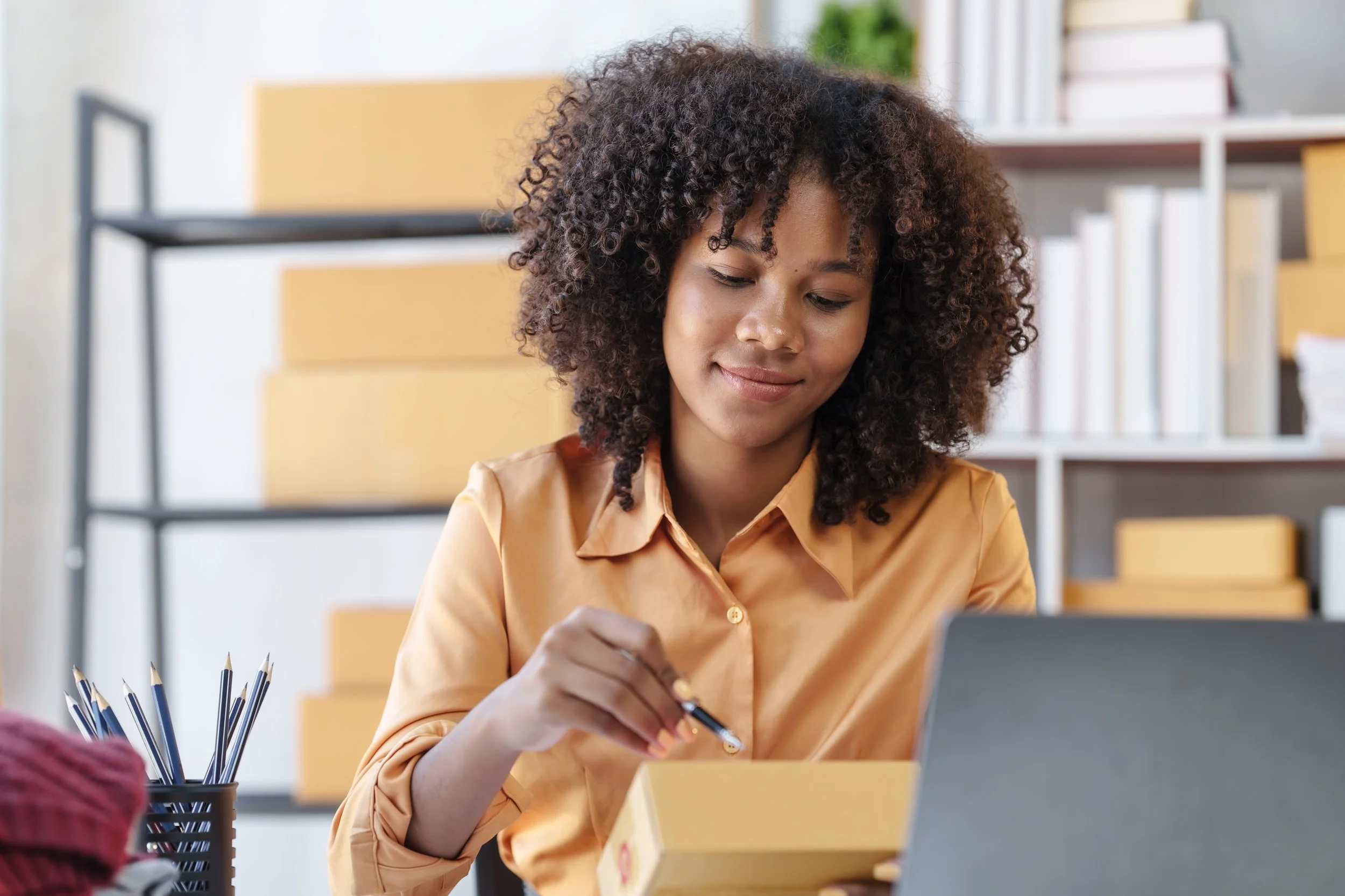 A young woman with curly hair looking down at a yellow box, with a laptop and a container of pens nearby in a modern office.