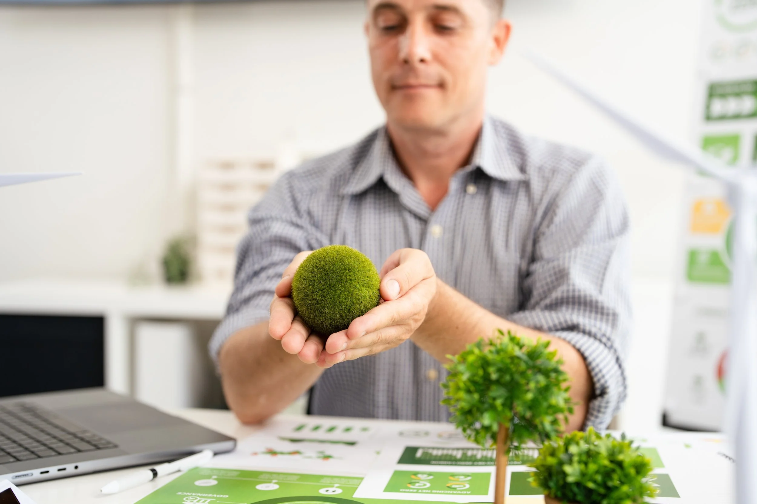 A man in a checkered shirt holding a green moss ball plant in a bright, modern environment, with papers and a laptop on the table in front of him.