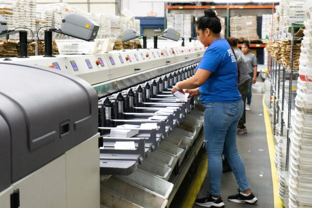 Women operating a paper shredder at a warehouse or mailroom, with shelves of packed boxes in the background.