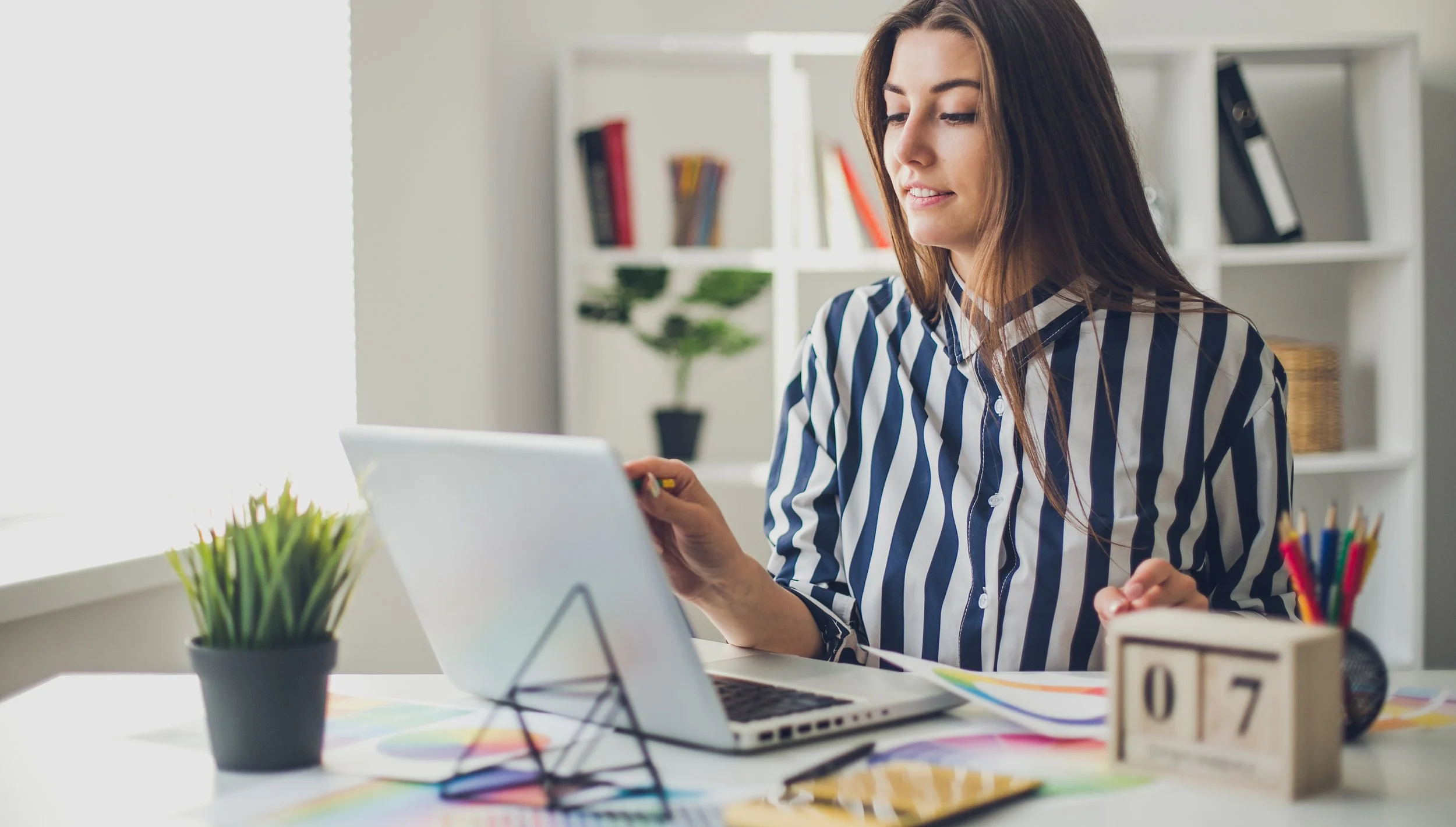 A woman sitting at a desk using a tablet, with a laptop, a potted plant, colored pencils, and a wooden calendar displaying 07 on the desk, in a bright room with bookshelves in the background.
