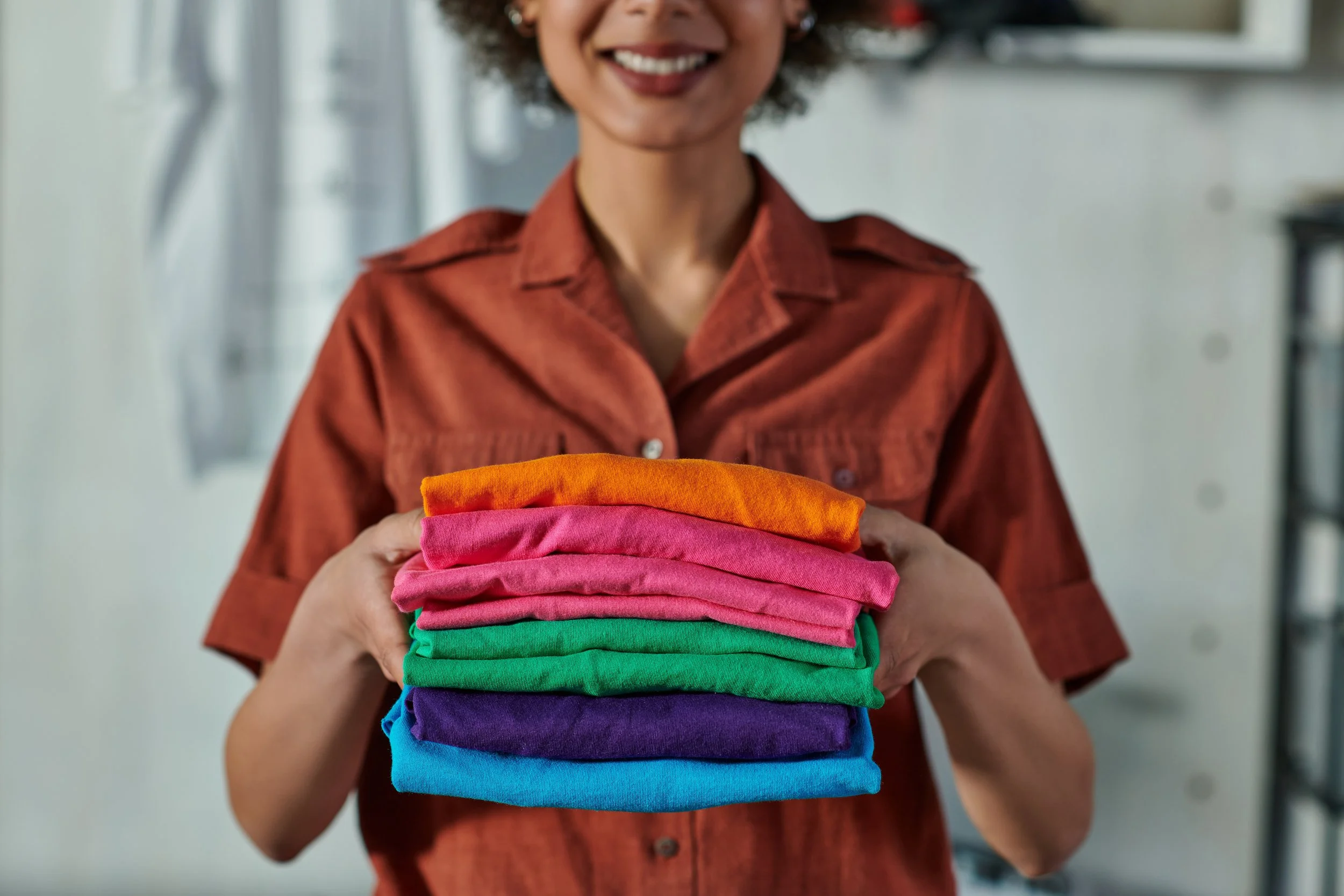Woman holding a stack of colorful folded shirts in a bright room.