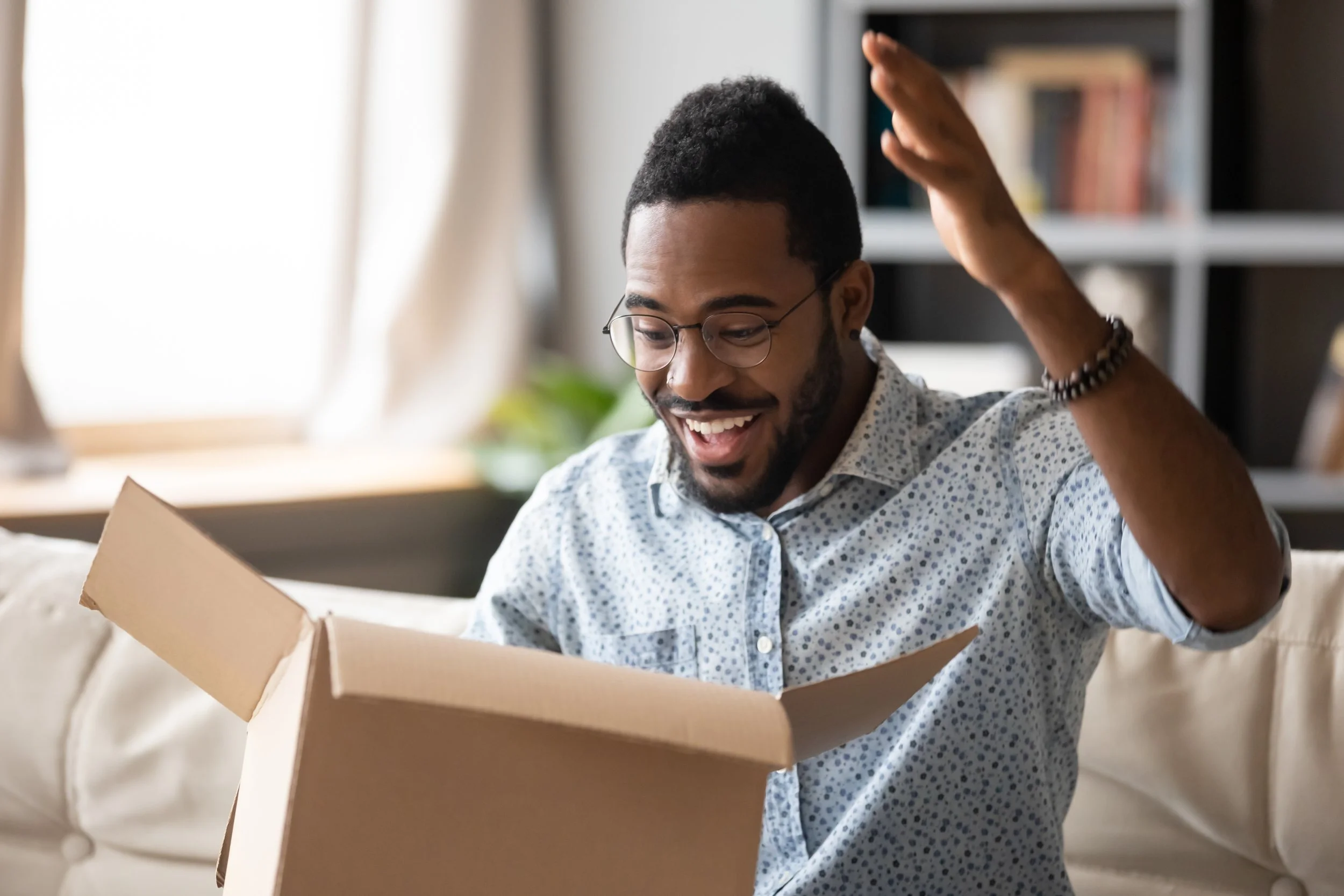 A man with glasses and a beard excitedly opening a cardboard box indoors, smiling with his hand raised.