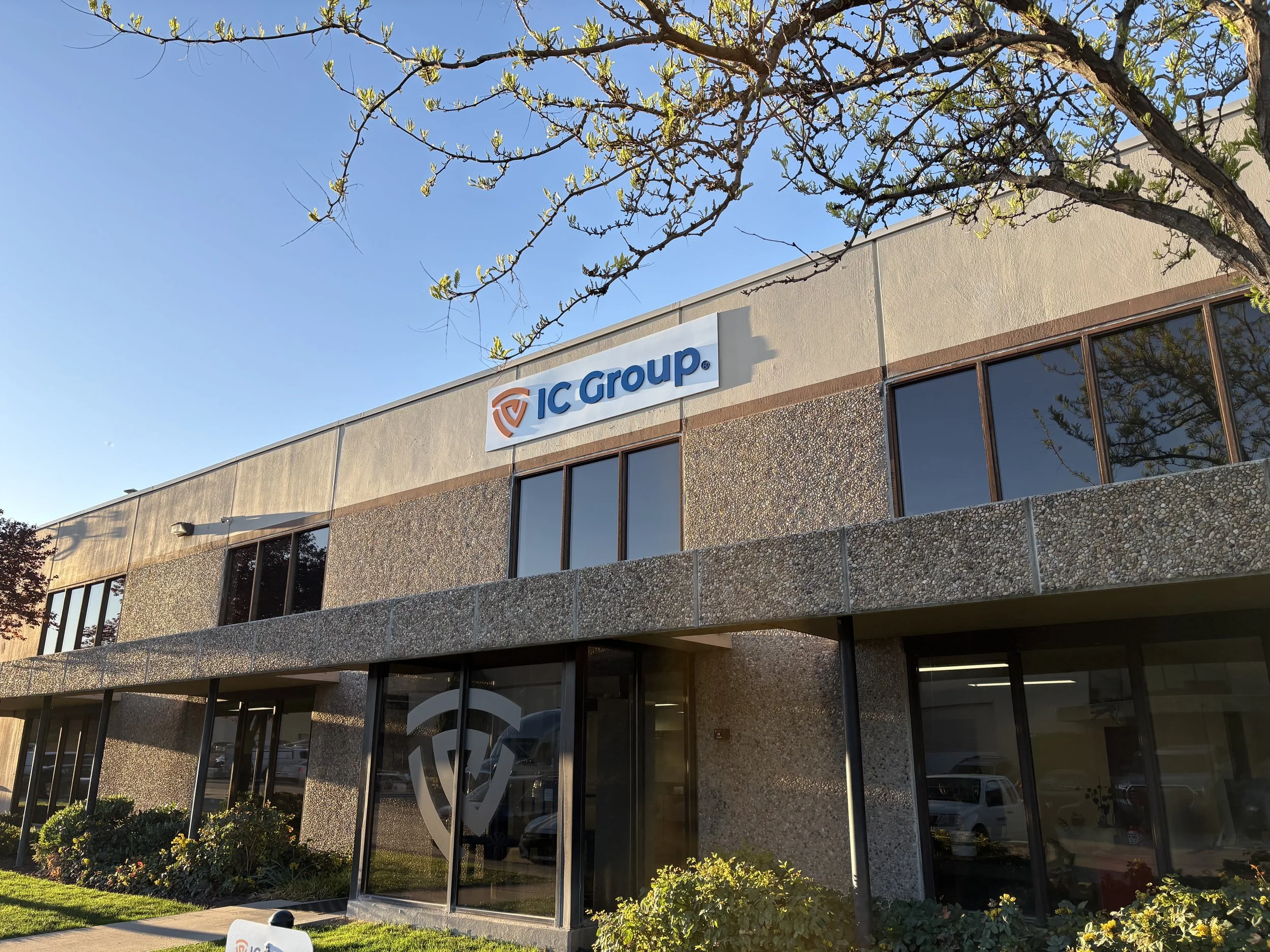 Exterior view of a two-story office building with a sign reading 'IC Group' and a logo on the upper facade. The building has a textured facade with large glass windows and a glass entry door. There are trees and bushes in front of the building, and the sky is clear with sunlight illuminating the scene.