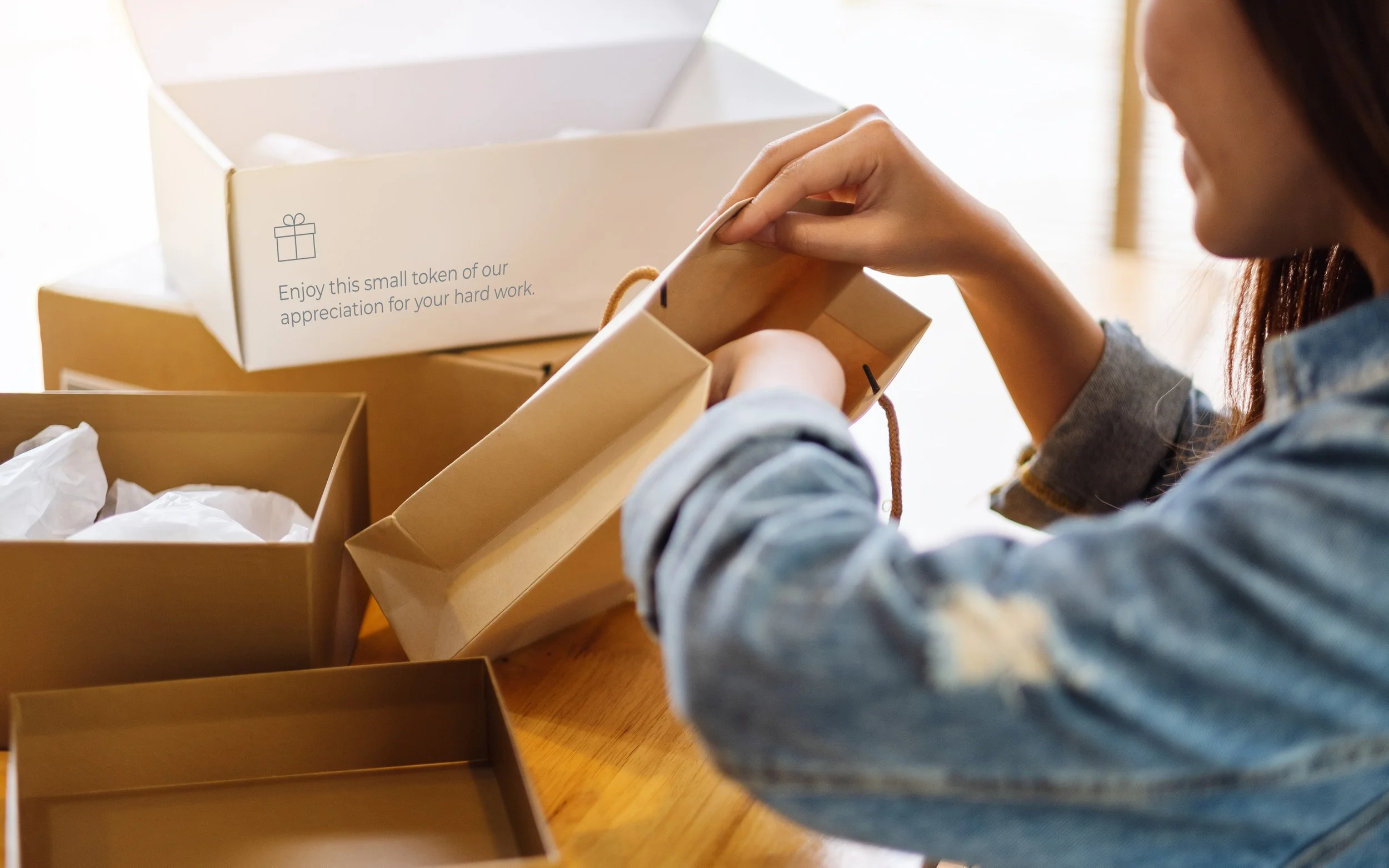 A woman in a denim jacket opening a small gift box with tissue paper inside, surrounded by larger boxes, one with a message about appreciation.