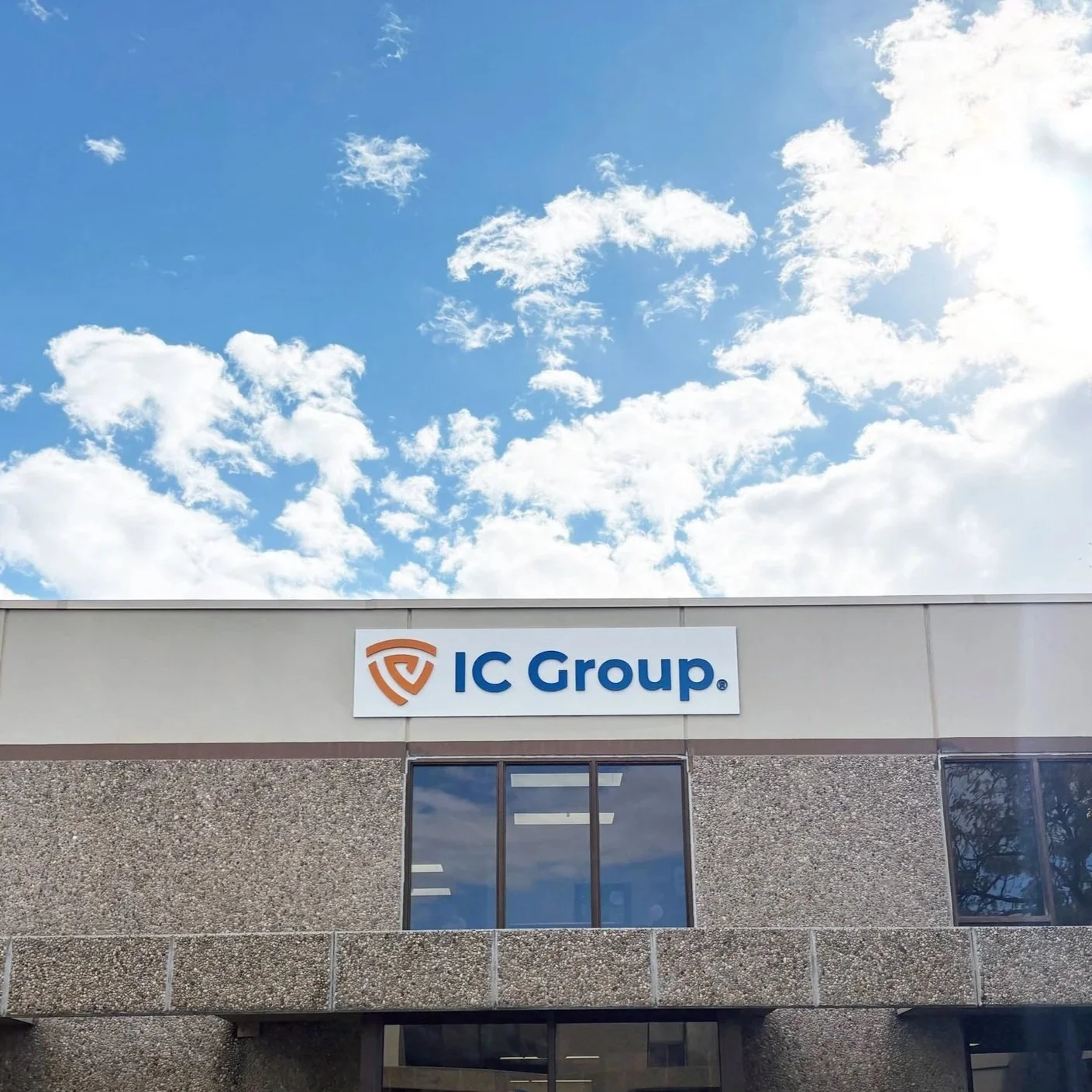 The image shows the exterior of a building with a sign that reads 'IC Group'. The sign is mounted above windows on the second floor of the building. The sky above is partly cloudy with a bright, blue sky.